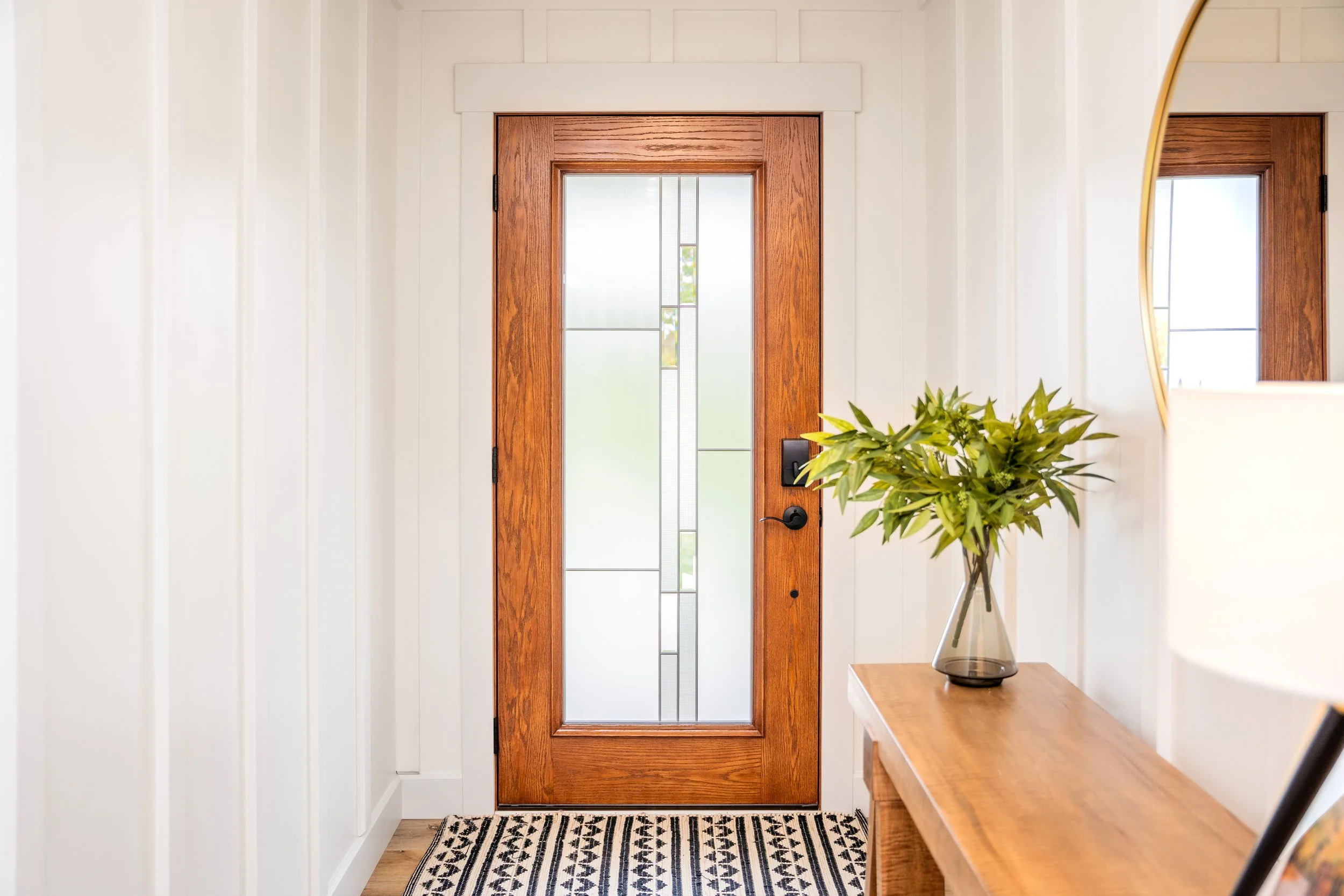 Interior view of a front door with frosted glass panels, a wooden frame, a black handle, and a lock, with a black and white patterned rug, a wooden console table holding a glass vase with green leaves, and a large mirror reflecting the door, in a wel