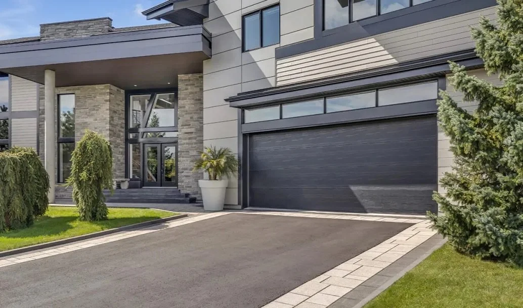 Modern house with stone and gray siding, large glass windows, black garage door, and neatly landscaped driveway with green bushes and trees.