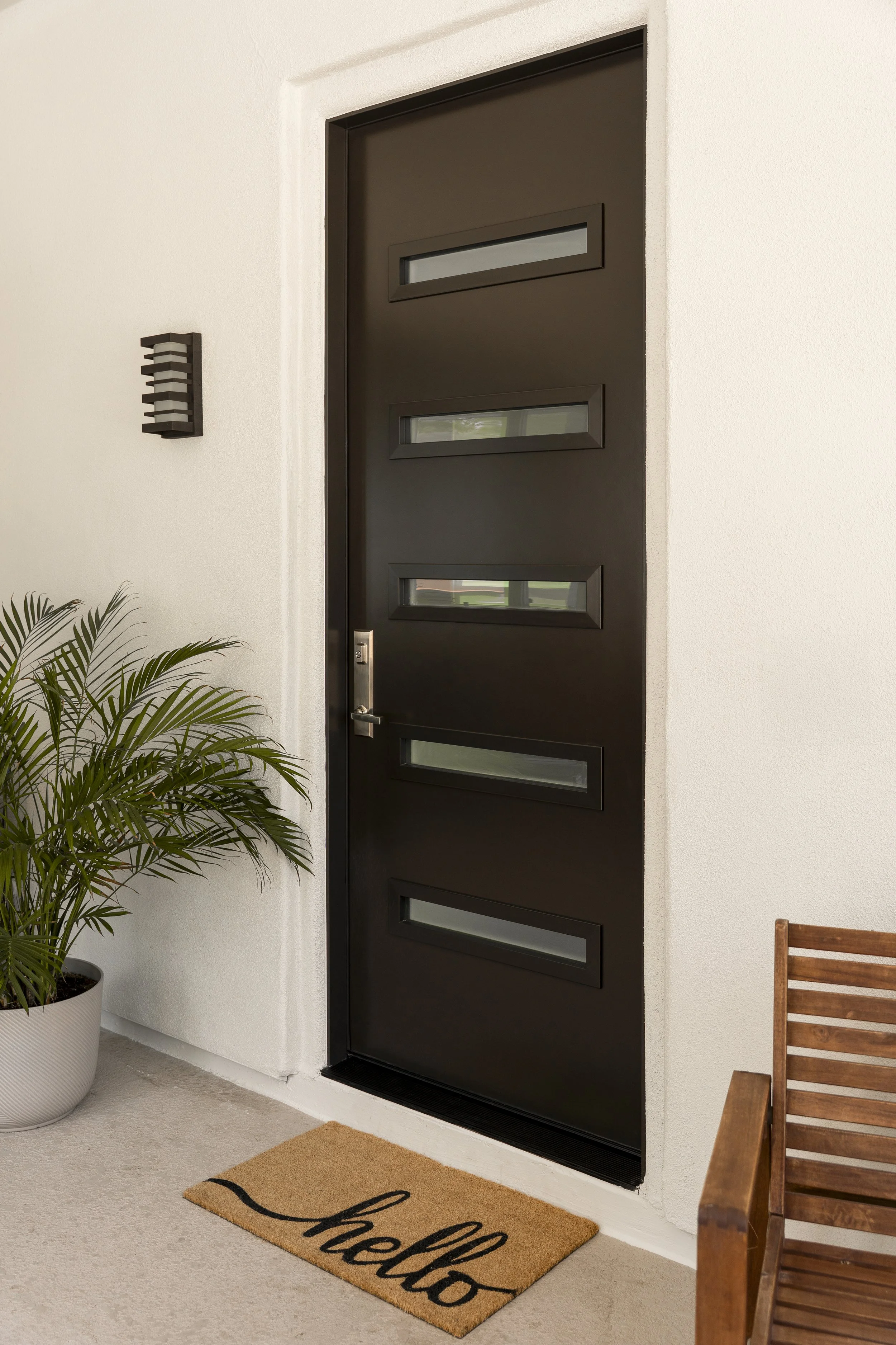 Black front door with four horizontal rectangular windows, white walls, potted plant, wooden chair, and 'hello' doormat.