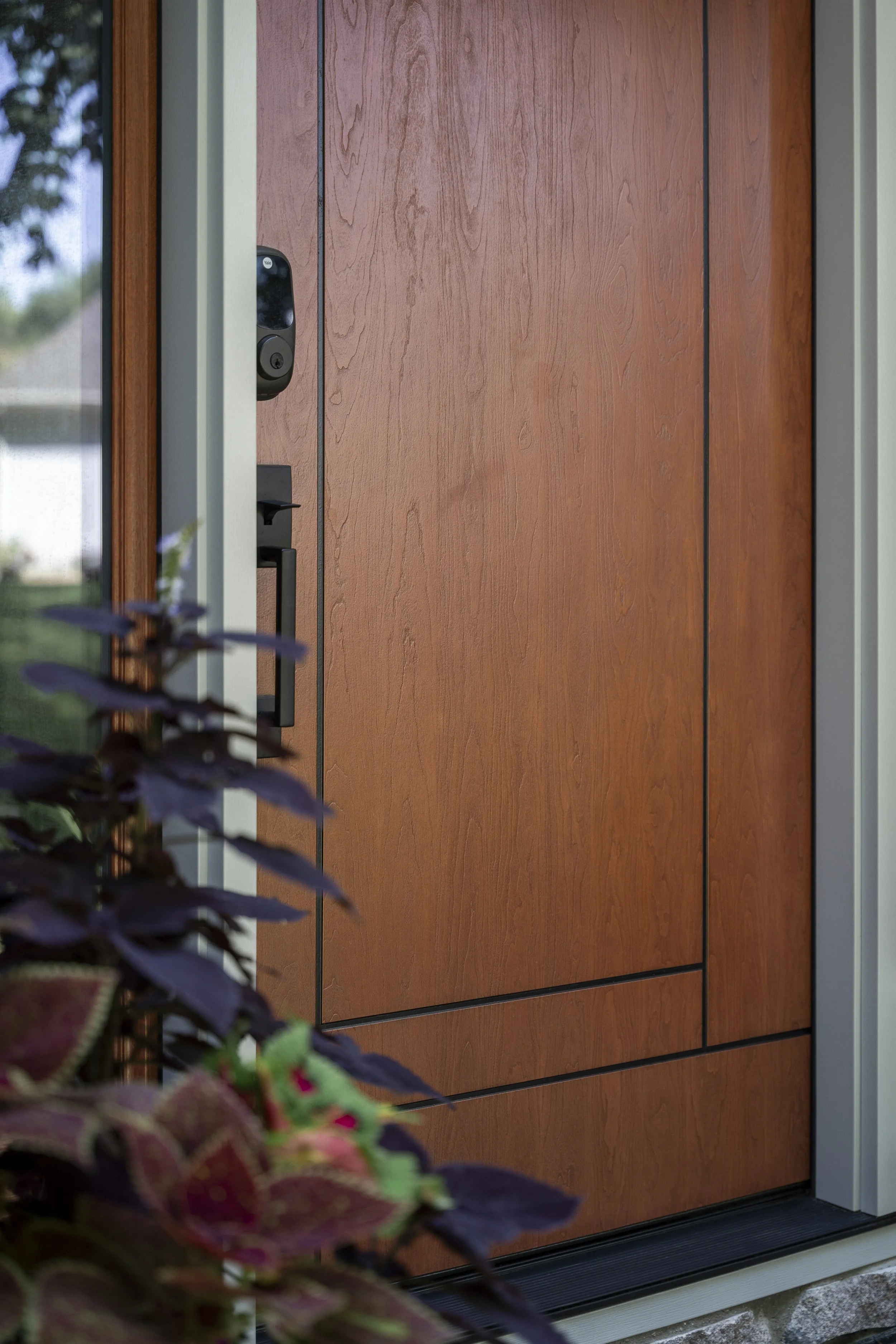 Close-up of a wooden front door with a black digital door lock and handle, partially obscured by purple and green plants.