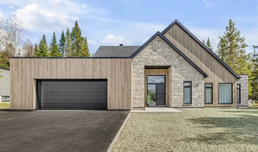 Modern house with a mix of wood and stone exterior, large black garage door, black front door, and tall windows, surrounded by trees and a cloudy sky.