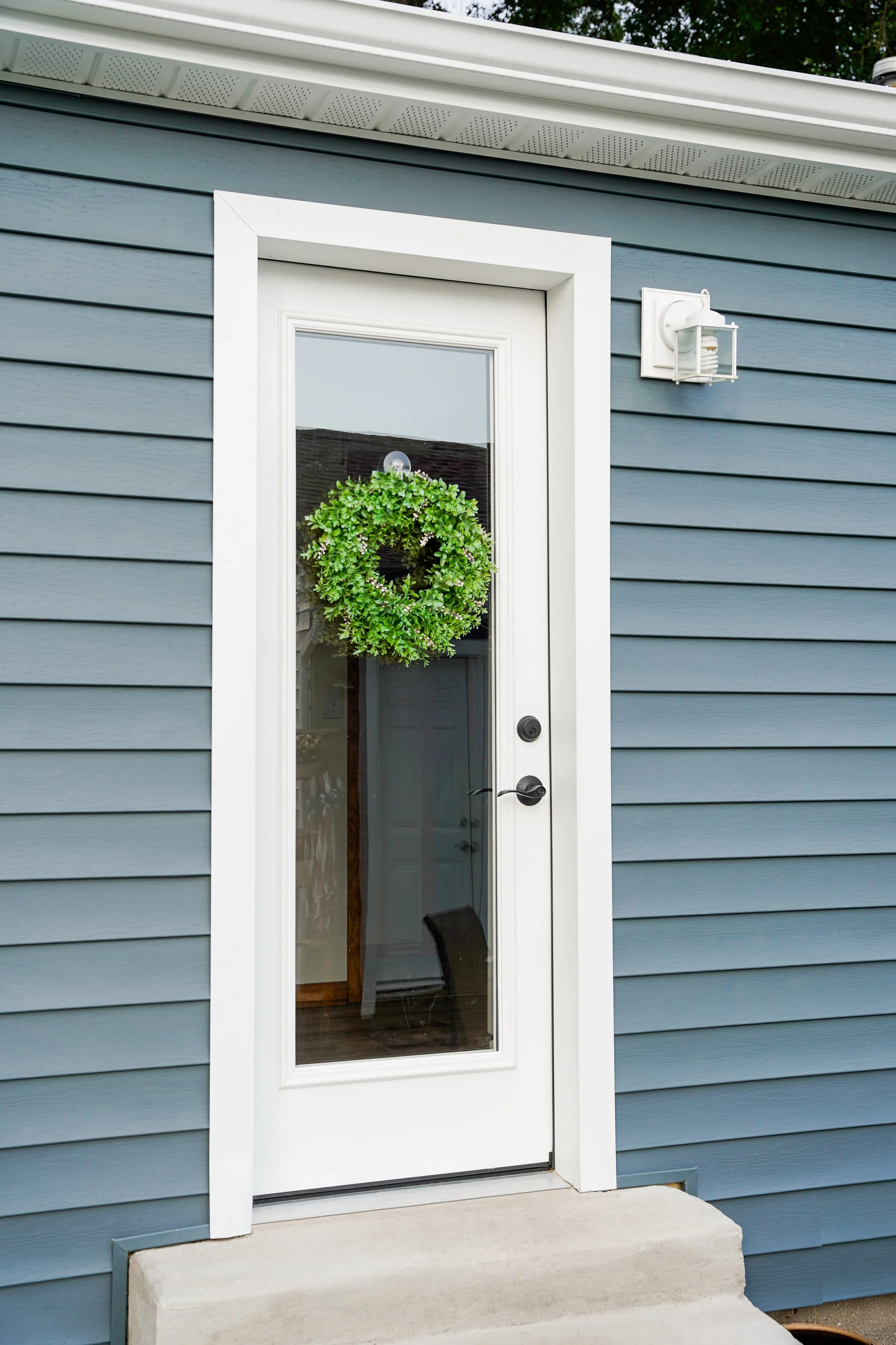 Blue house exterior with a white-framed glass door decorated with a green wreath, beside a white outdoor wall lantern.