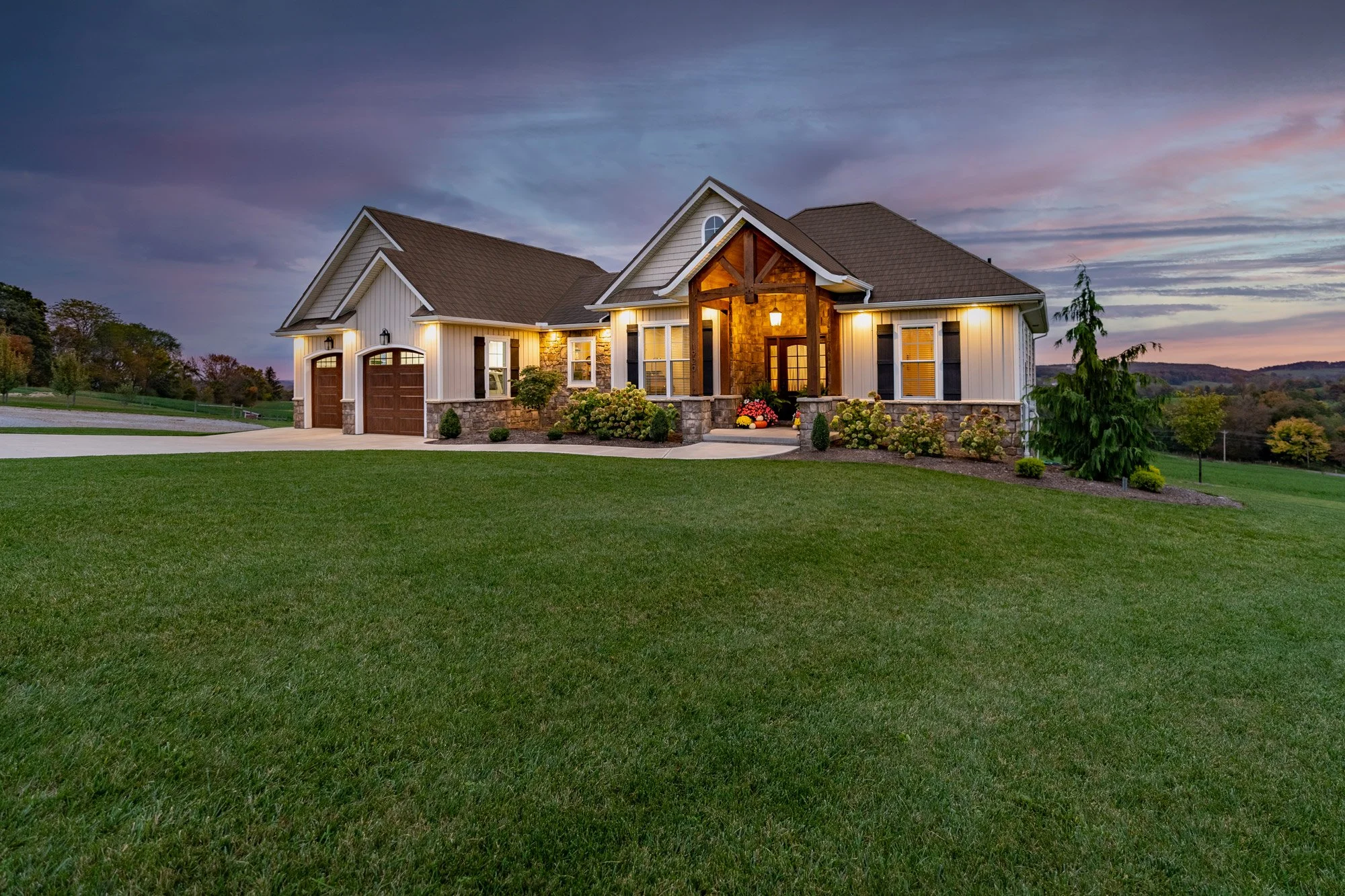 A large, modern house with a landscaped front yard and a driveway, illuminated by exterior lights at dusk.