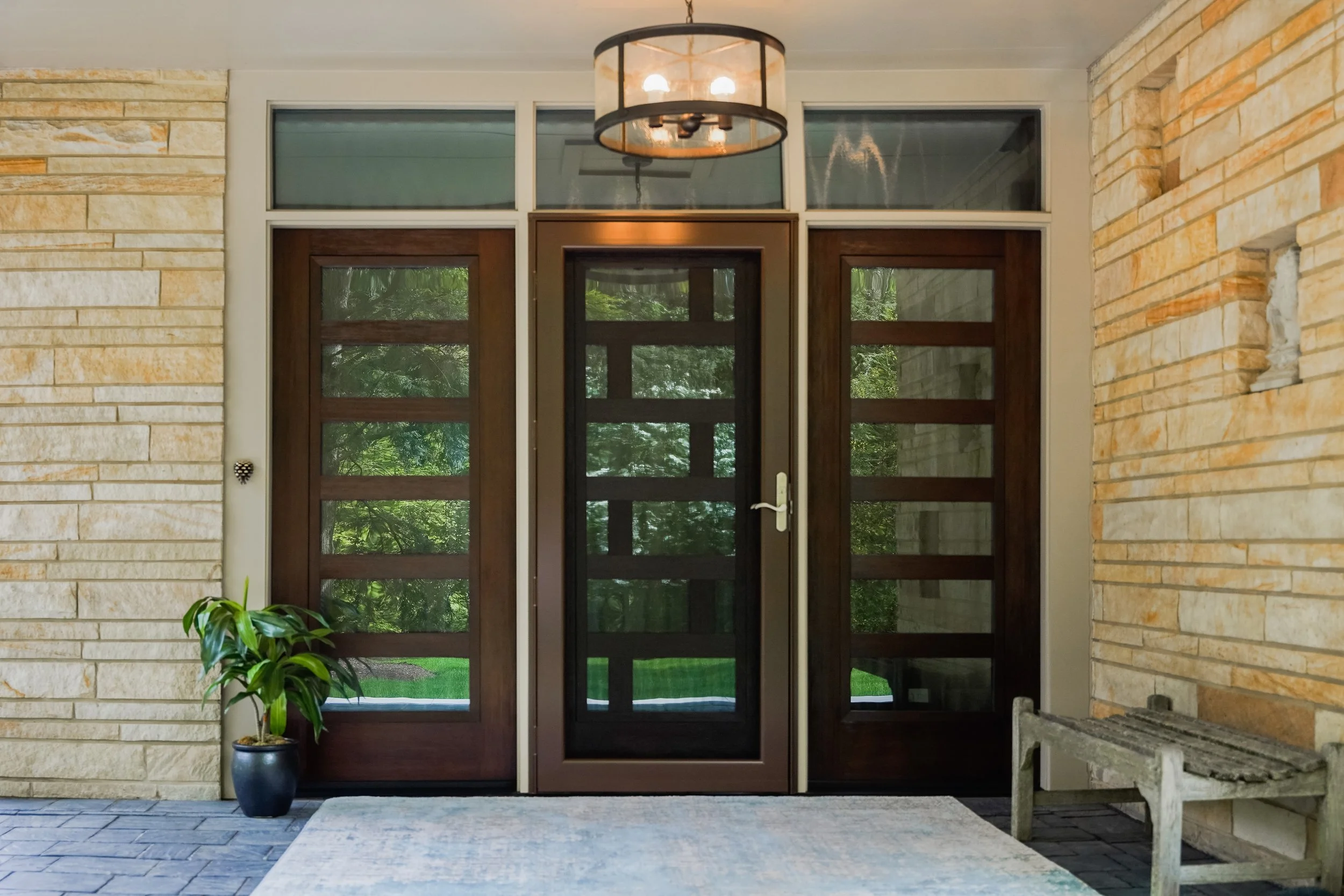 Modern front door entrance with glass-paneled double doors and a central screen door, surrounded by stone walls, a potted plant on the left, a small wooden bench on the right, and a ceiling light fixture, with lush greenery visible through the glass.