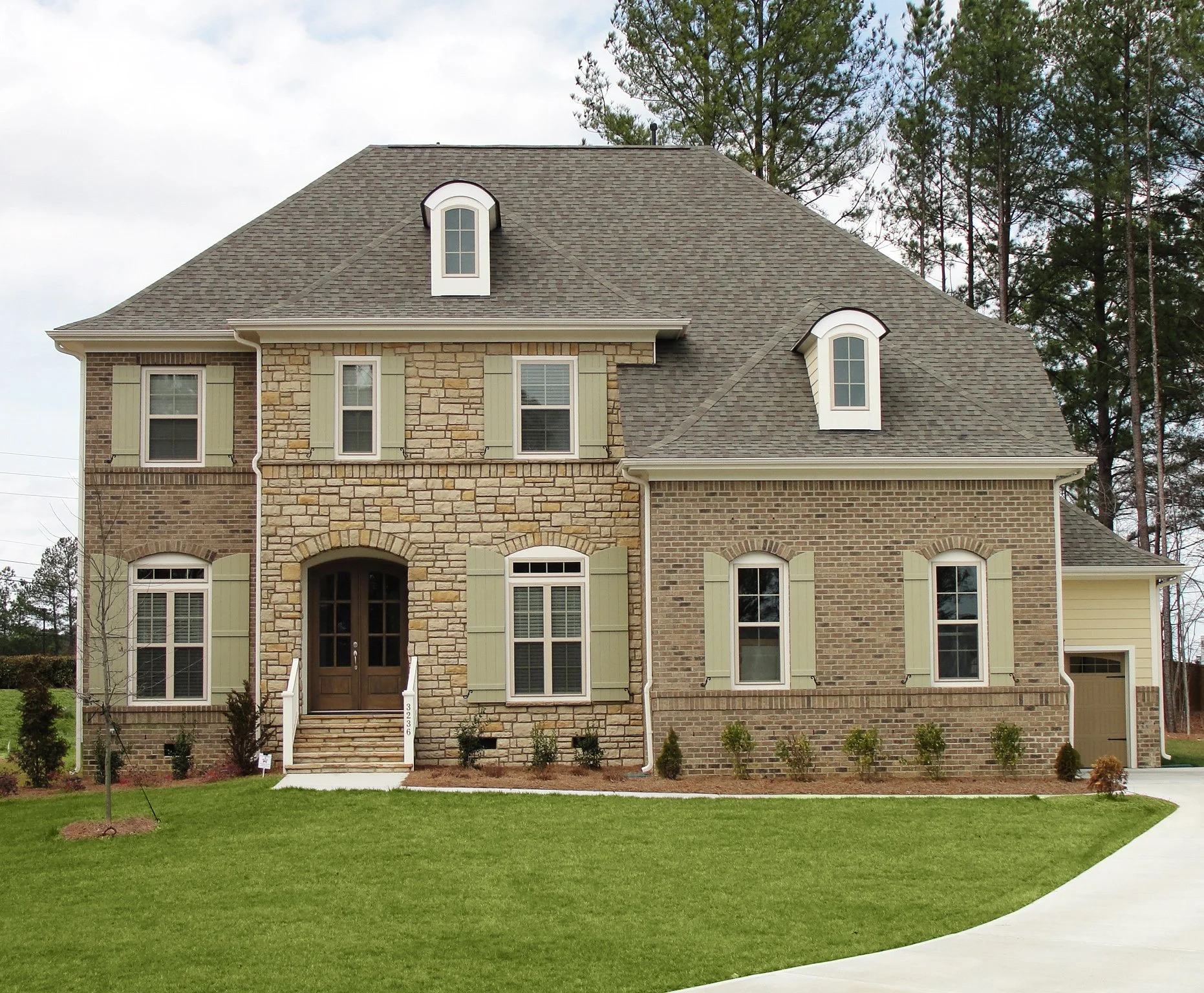 Front view of a large two-story house with brick and stone exterior, multiple windows with light green shutters, a gabled roof with dormer windows, and a well-maintained green lawn with small bushes and a sidewalk.