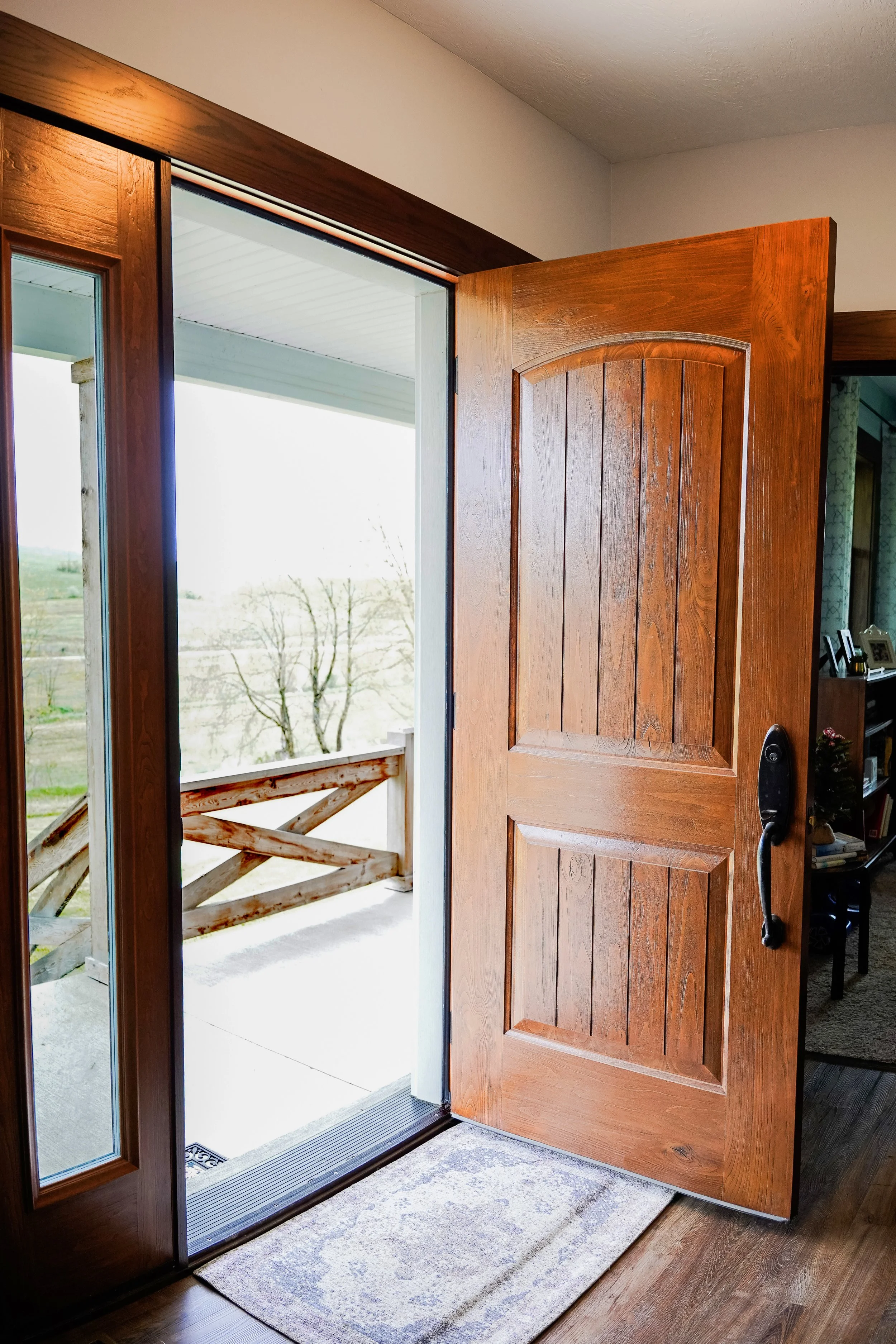 Open wooden front door leading to a porch with a view of a rural landscape and trees in the distance.