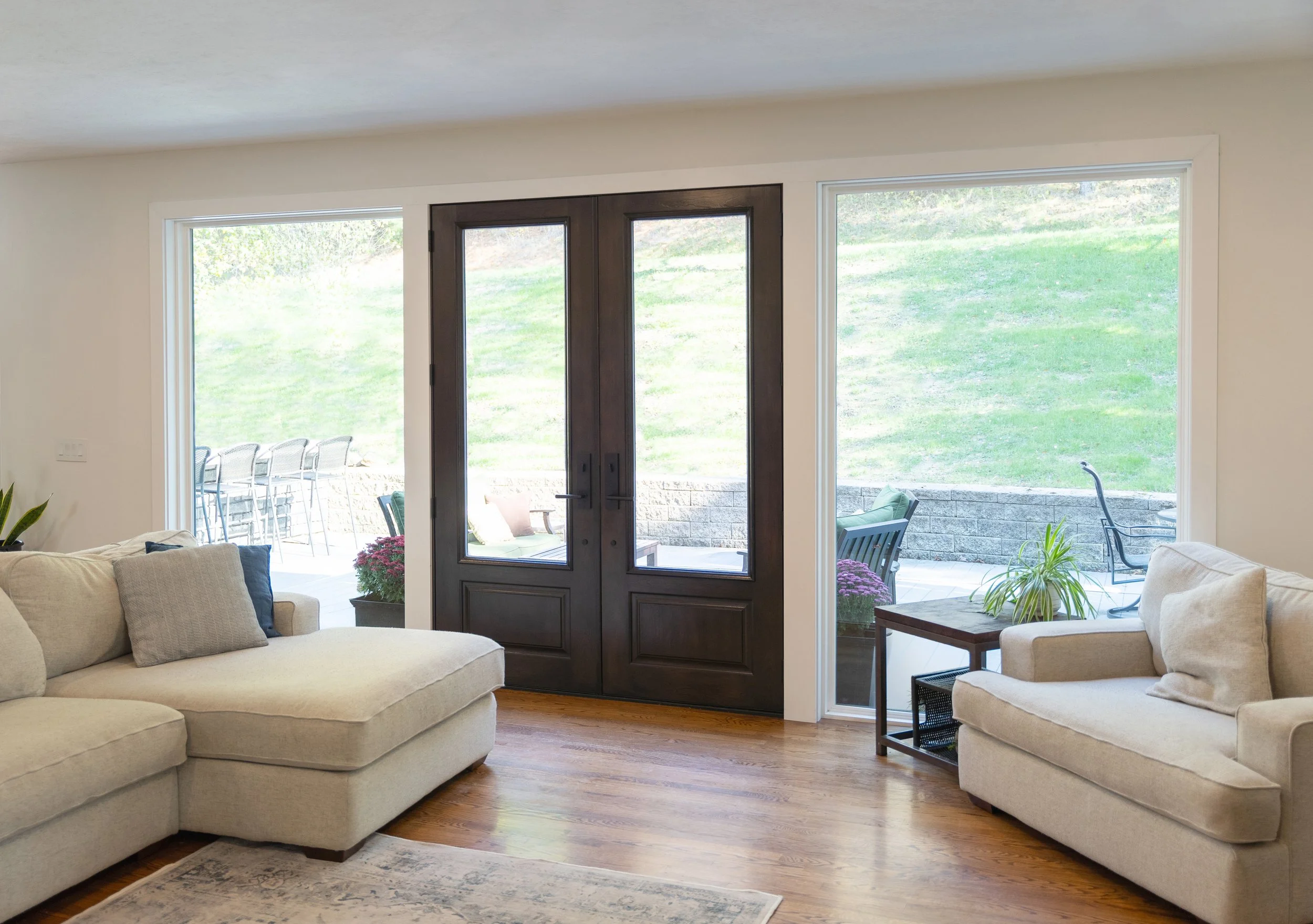 Living room with beige sofas, hardwood floor, dark wood double front door, large windows showing outdoor patio and grassy yard, potted plants, and side table.