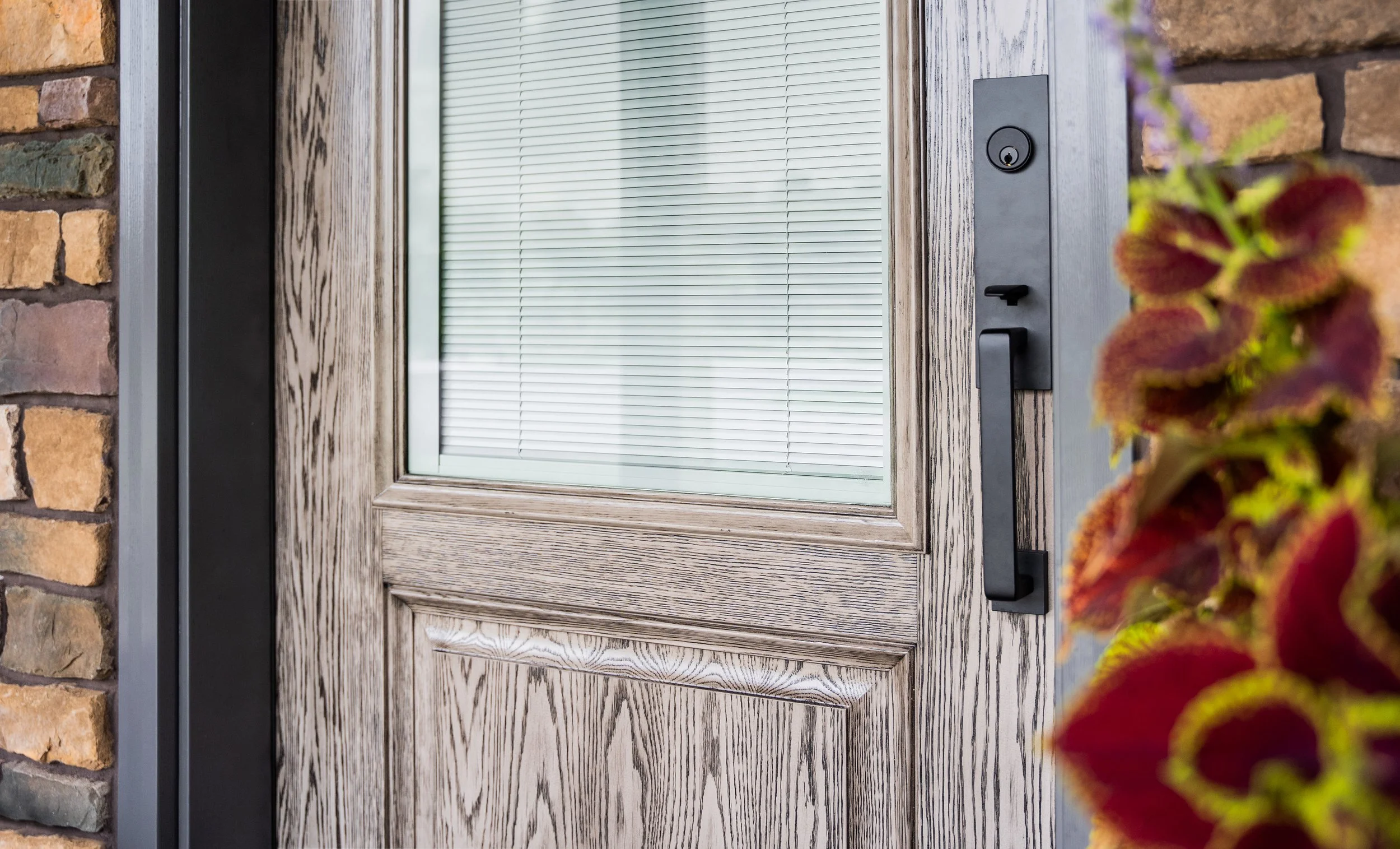 Close-up of a wooden front door with a small window and black handle, part of a brick wall with a plant with red and green leaves visible on the right.