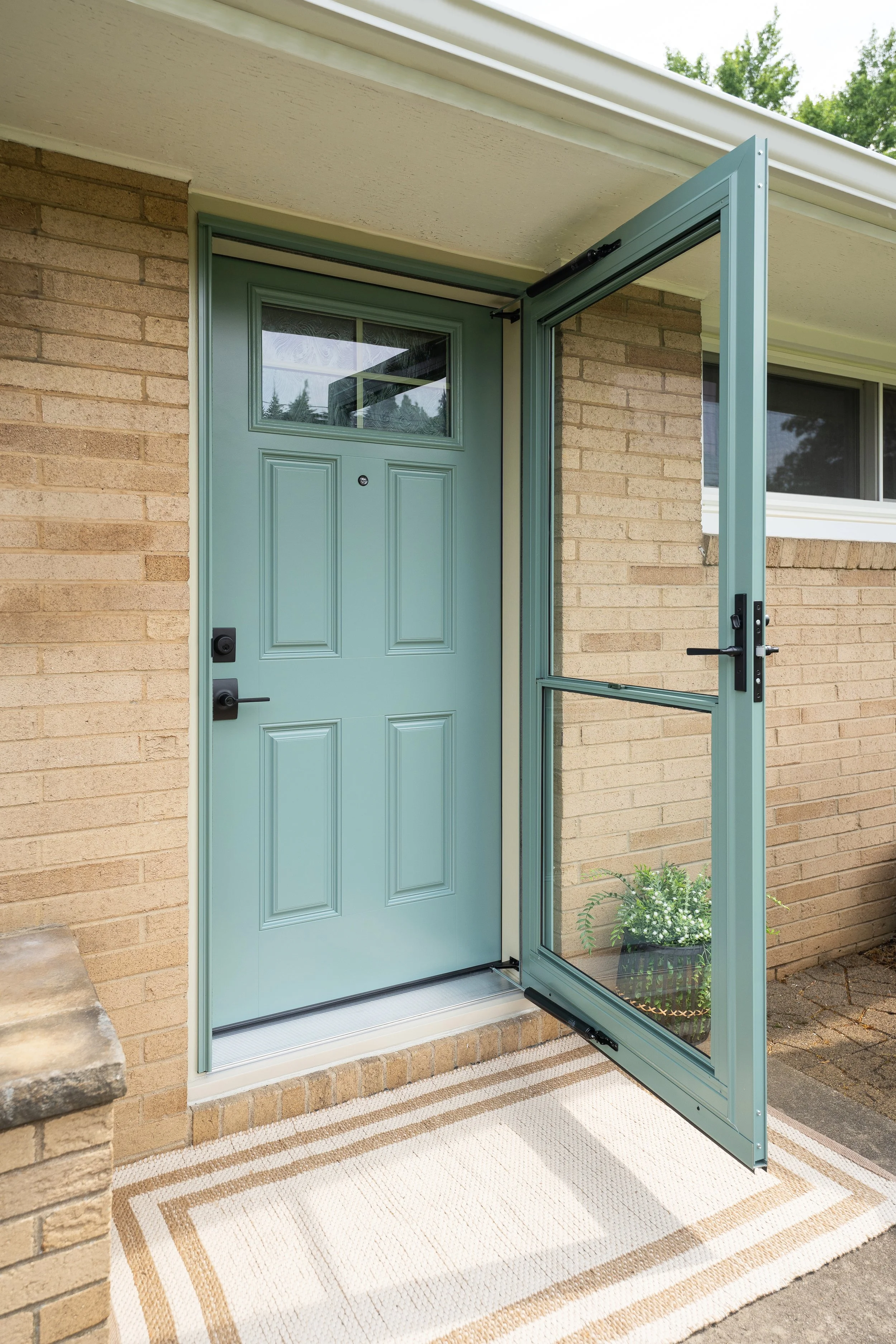 A light blue front door with a small window at the top, surrounded by a gray frame. The door is slightly open, revealing a screen door. There is a beige and white striped welcome mat in front of the door, and a potted plant visible through the screen