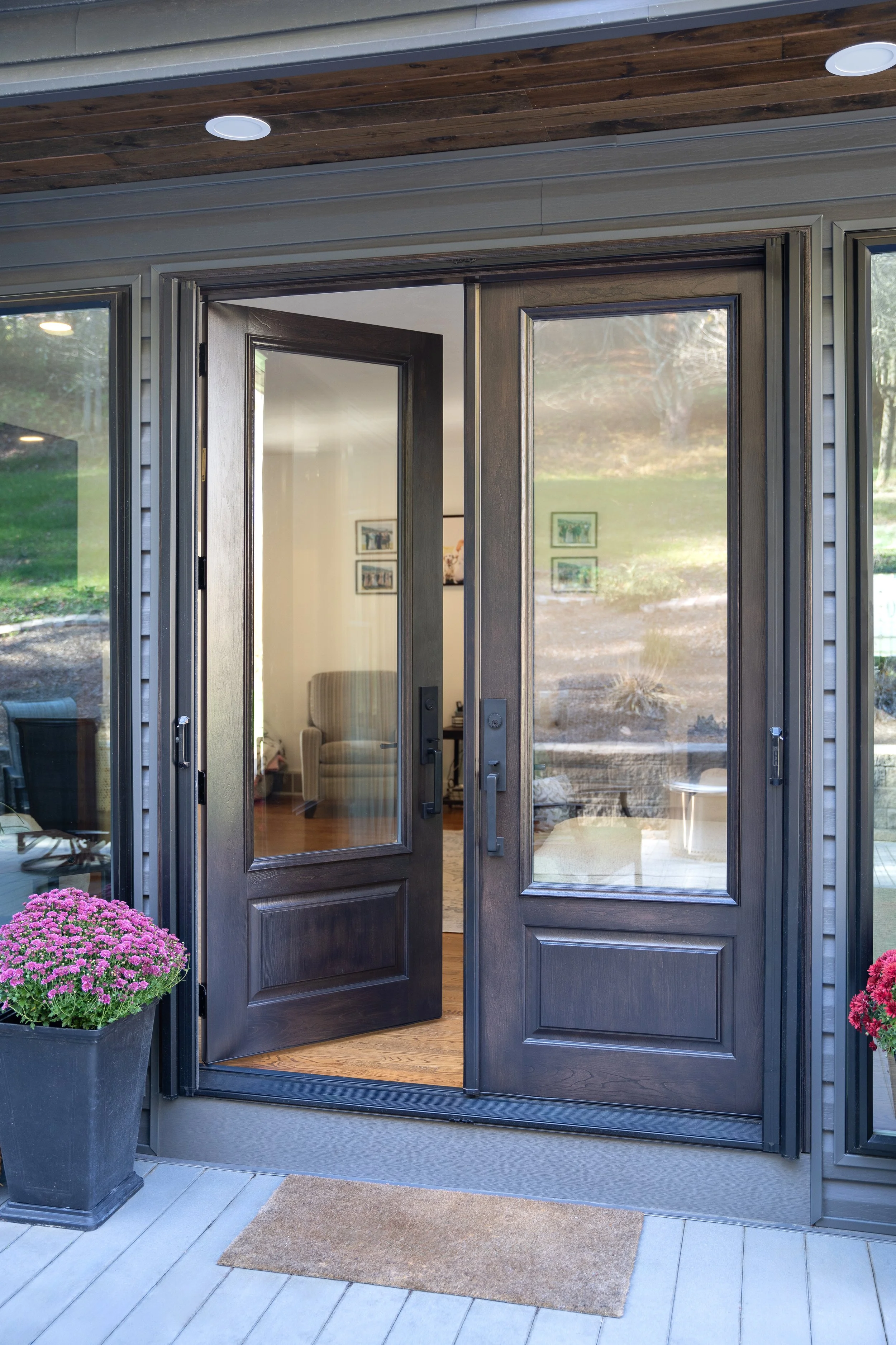 A front porch with black glass patio doors, one door open, revealing a living room with an armchair and framed pictures on the wall inside. Two potted pink flowers are on either side of the doors, and a doormat is on the porch floor.
