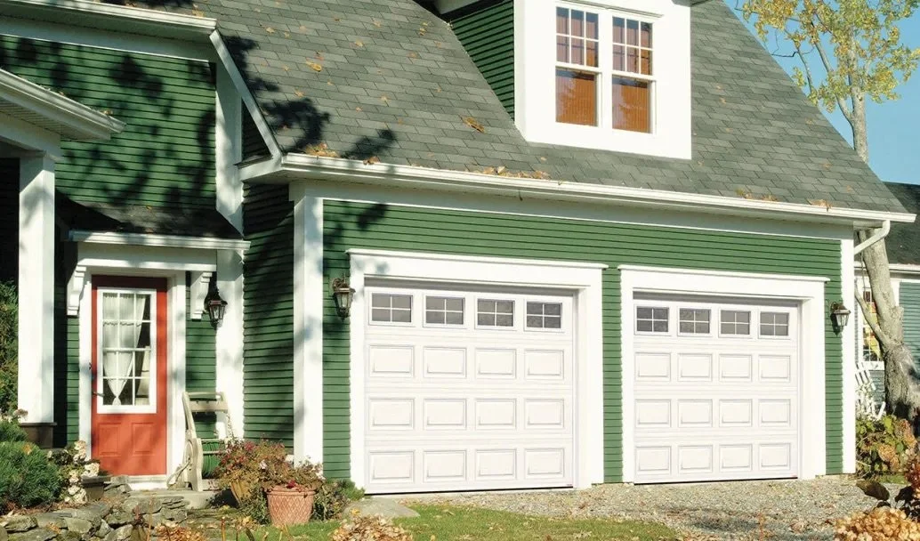 Front view of a green house with white trim, featuring a red front door and two white garage doors, with a tree on the right side.