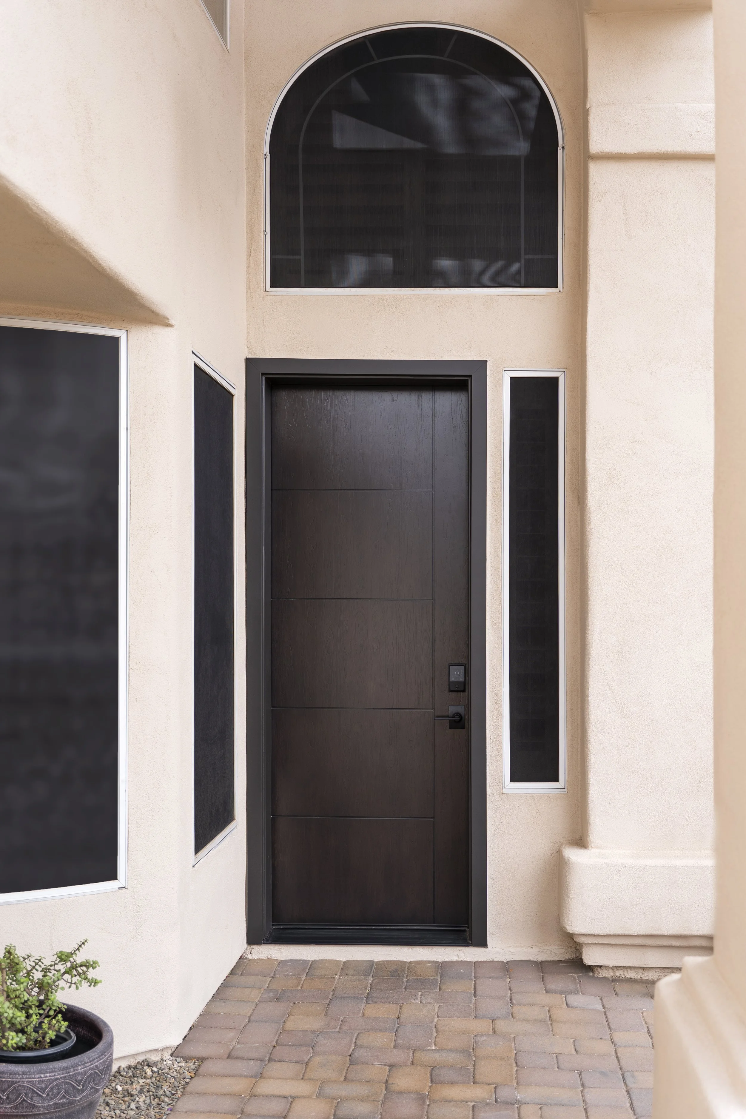 A modern front door with a black finish, surrounded by beige stucco walls with rectangular black windows and a semi-circular window above, on a paved brick porch.