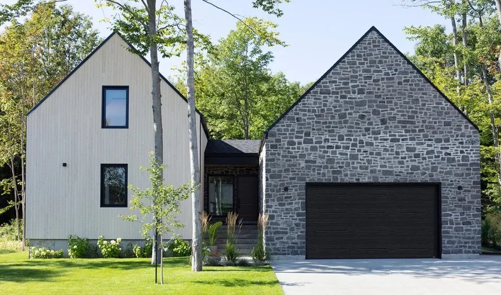 Modern house with two distinct sections: one with white vertical siding and one with gray stone facade, a garage door, and a front yard with grass and small trees.