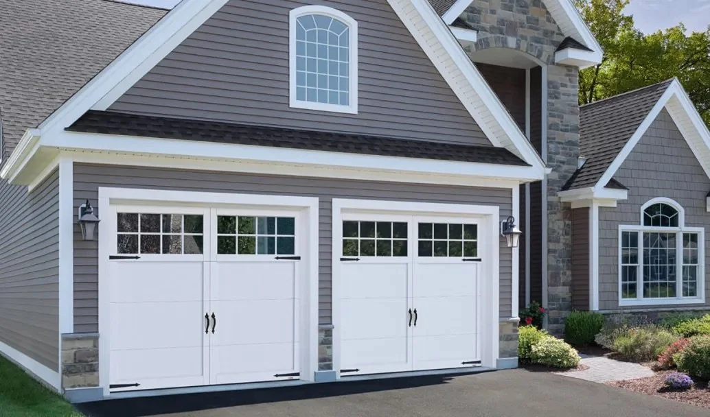 A modern house with gray siding, white trim, and two white garage doors with windows at the top. The house features stone accents and large windows, surrounded by a landscaped yard with bushes and flowers.
