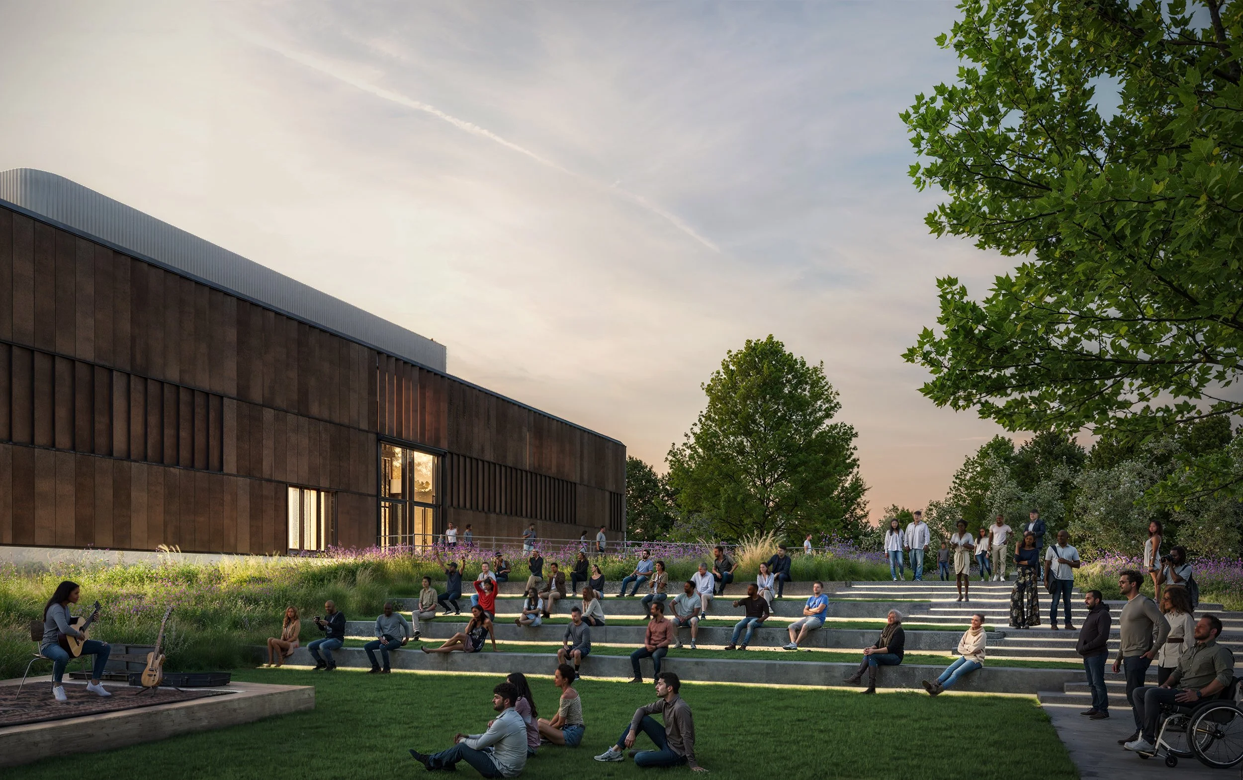 People gather on outdoor tiered steps and lawn, some sitting and others standing, while a woman plays guitar on a small stage at sunset, trees and a modern building are in the background. 
