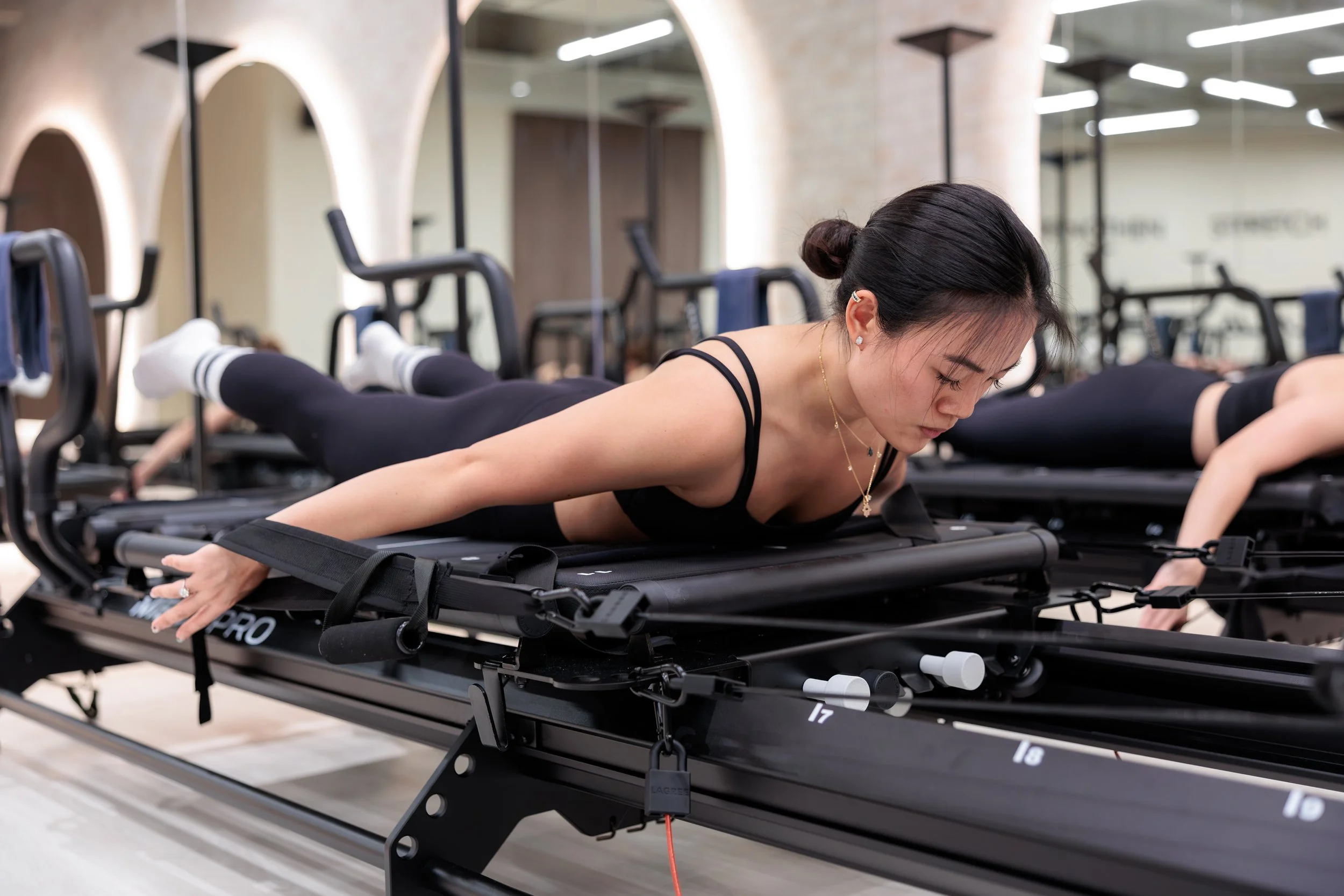 A woman is doing reformer Pilates on a Pilates reformer machine in a gym, with another person visible nearby. HKore Megaformer Lagree Yoga Studio Photography