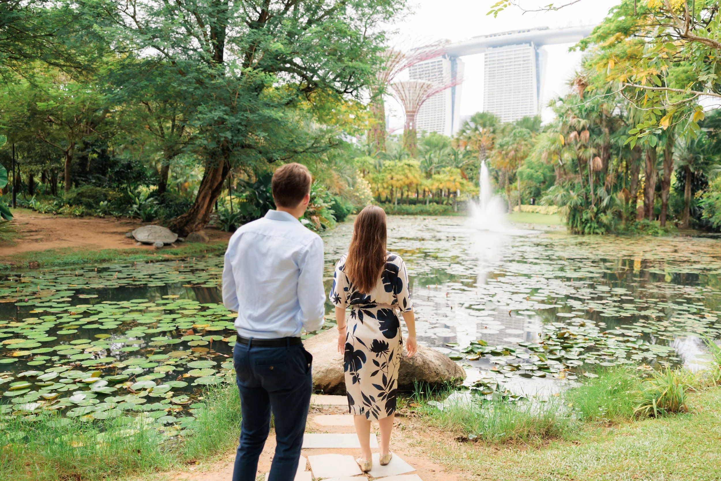 Two people walk towards a pond with a fountain, surrounded by lush trees and greenery, with modern high-rise buildings visible in the background. Singapore Proposal Photographer waiting for Surprise Proposal.