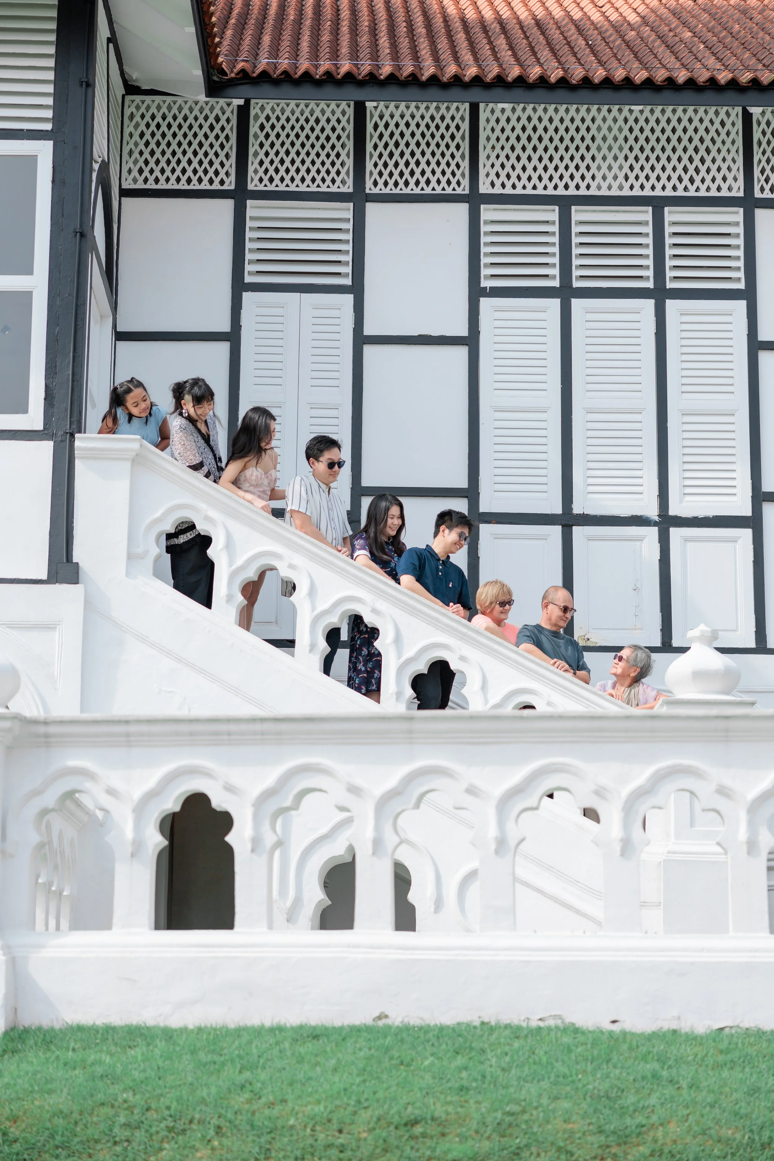Group of people walking down a white decorative staircase outside a house with dark wood trim and a red tiled roof. Singapore Outdoor Family Photography. Singapore Family Photography.