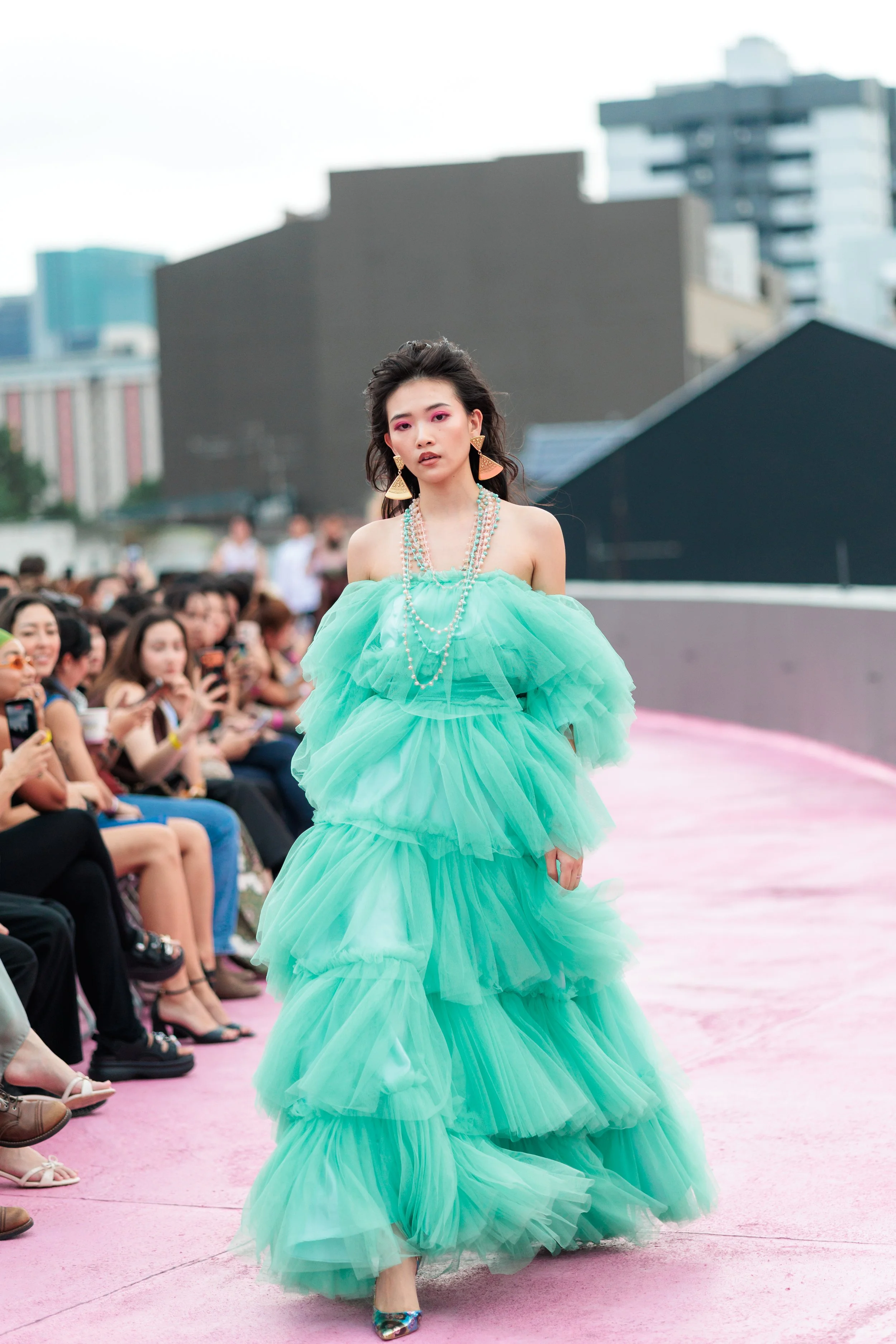 Model walking on pink runway during fashion show, wearing a vibrant, layered, mint green ball gown with off-shoulder design, multiple pearl necklaces, large triangle earrings, and colorful high heels, with an audience seated on one side and an urban 