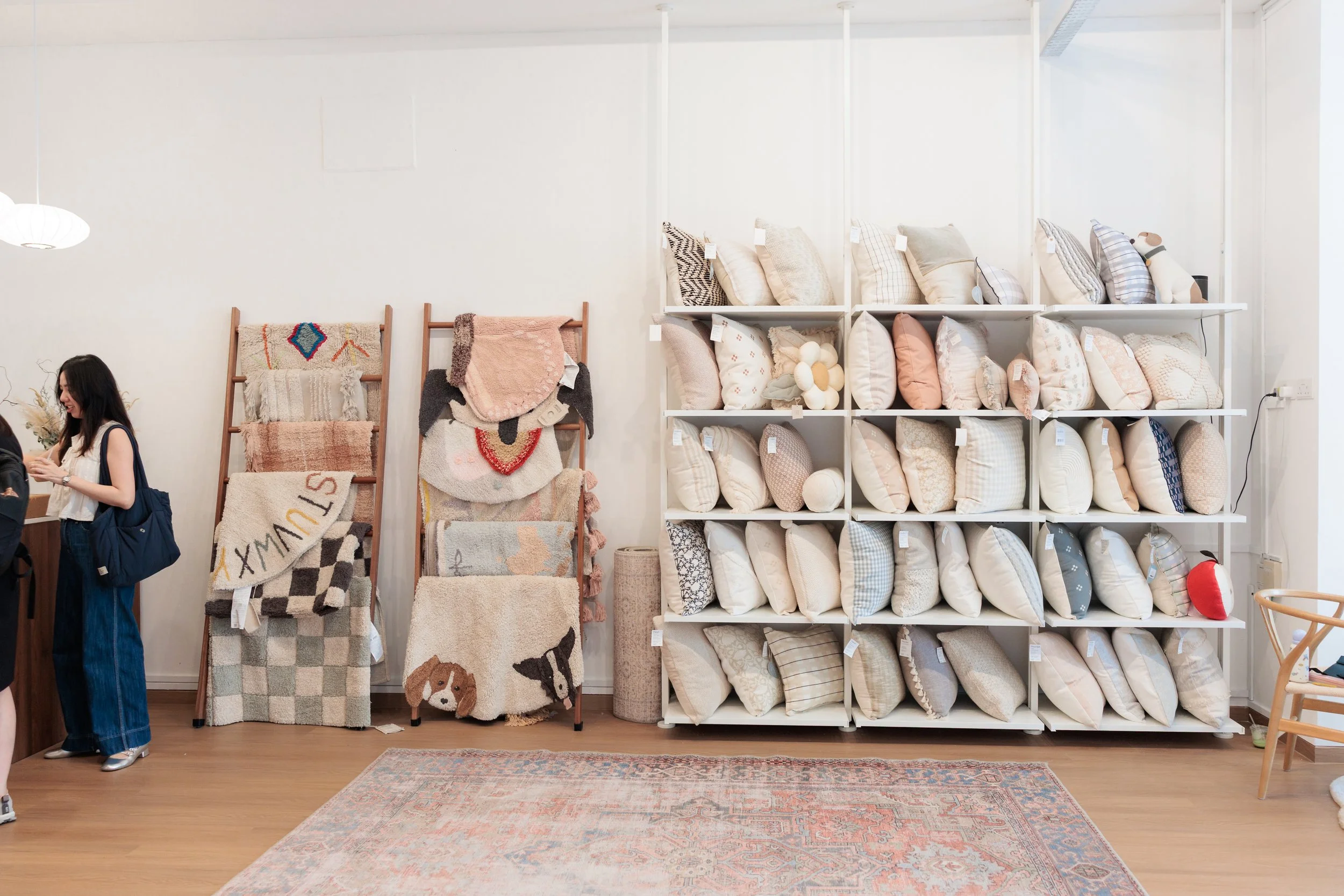 A display of pillows on white shelves, with decorative rugs hanging on stands and a person with a dark backpack on the left side of the image in a home decor store. Loom & Stone Store Opening. Event Photography. Event Videography. Rugs. Carpets. Mari
