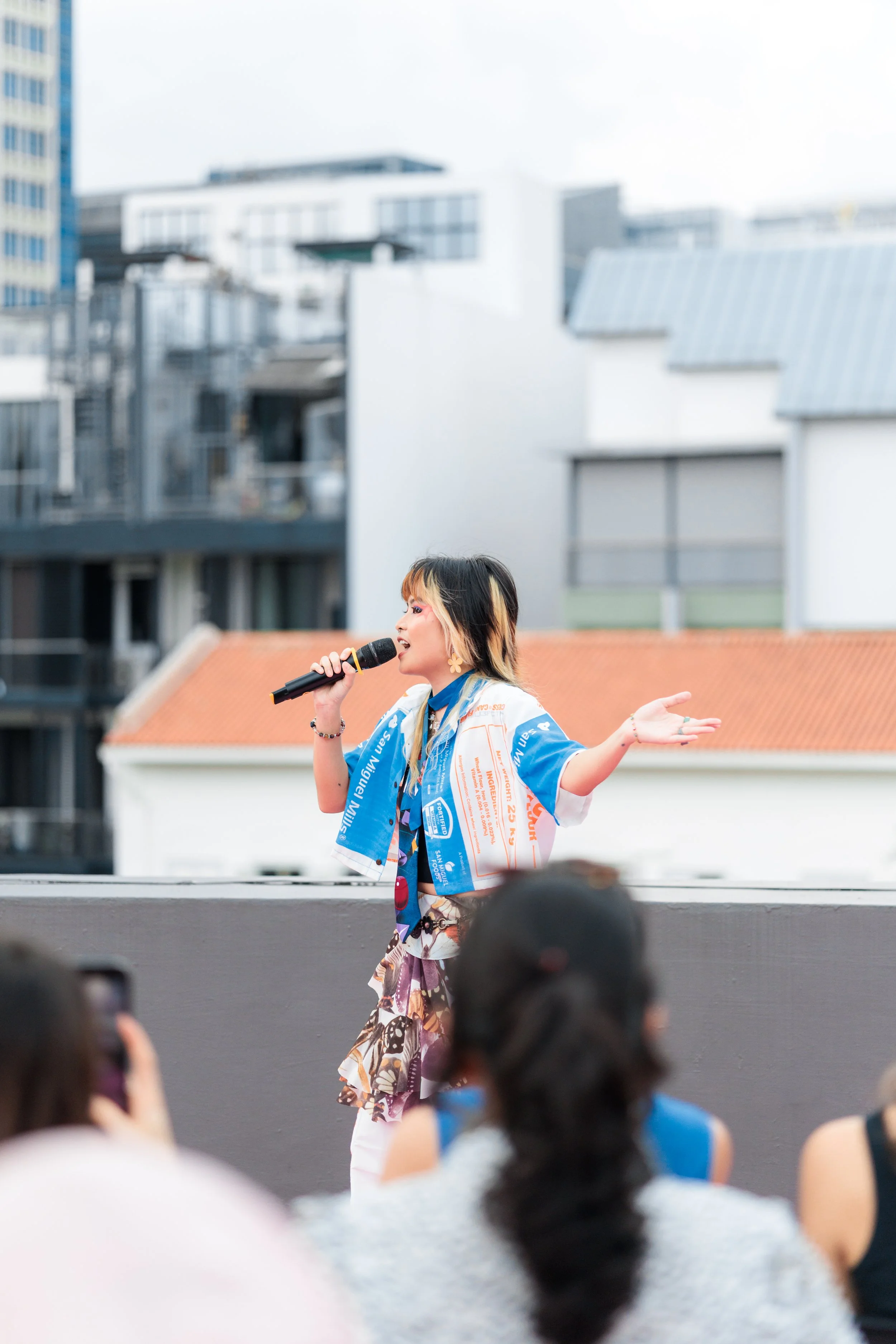 A woman with multi-colored hair and earrings singing during an outdoor event, with a crowd of people in the foreground and buildings in the background. Event Photography. Fashion Photography. Event Videography. Walkway Photography & Videography.