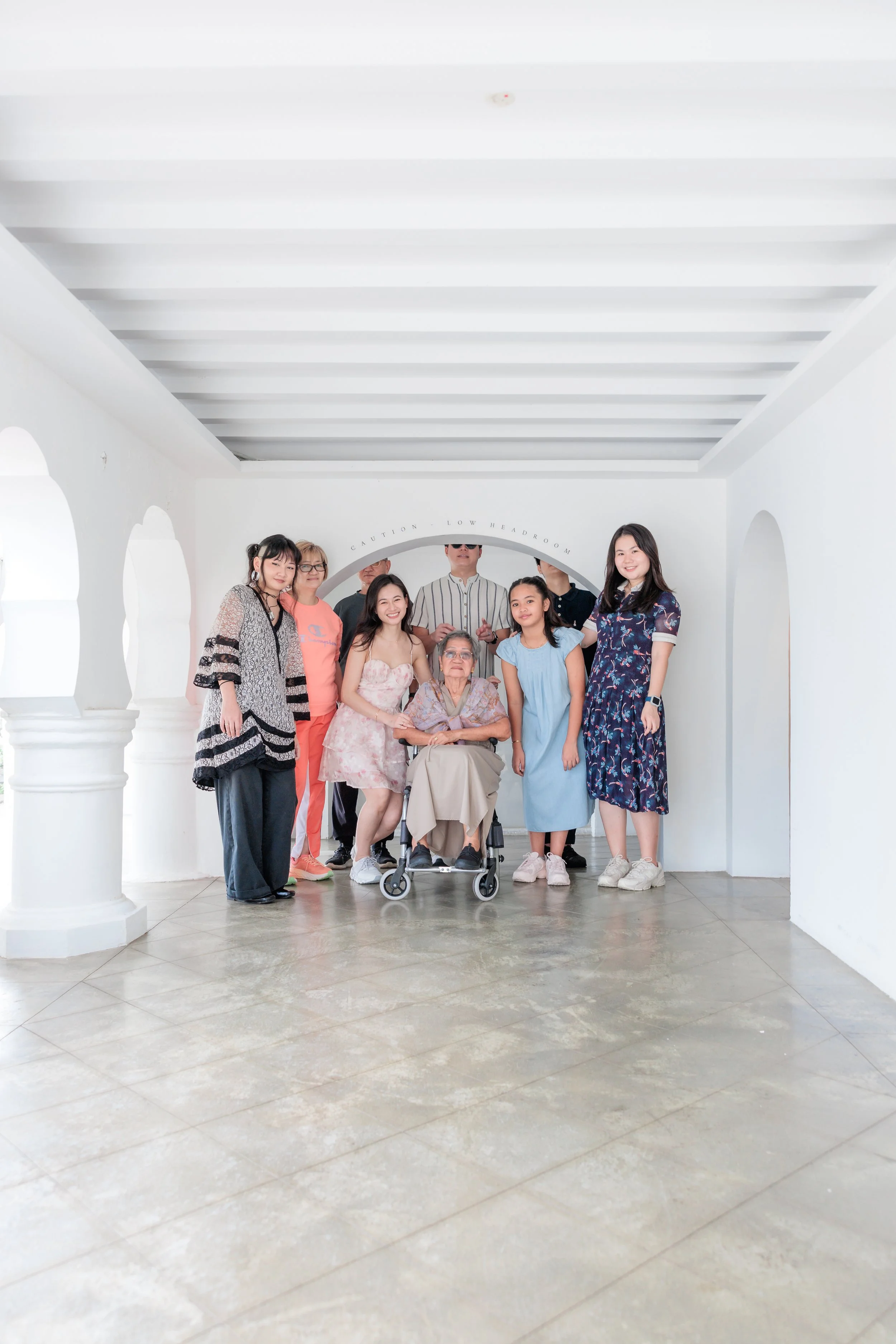 A diverse group of nine people, including an elderly woman in a wheelchair, posing inside a white building with arches and a ceiling with exposed beams. Singapore Outdoor Family Photography. Singapore Family Photography.