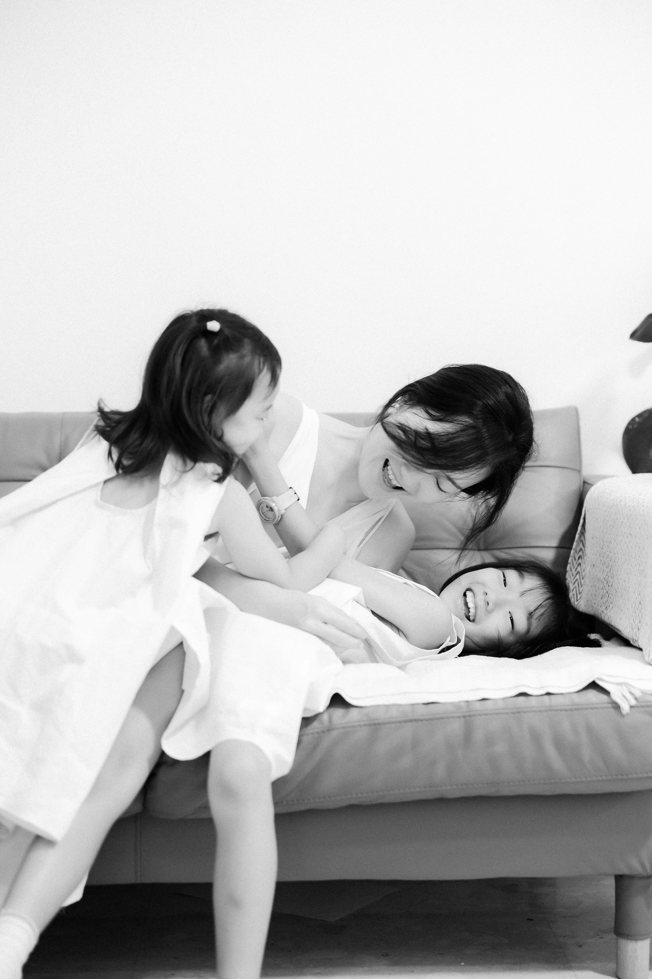 A woman and two young girls playing and laughing on a couch in a living room. Family Photography. Family Photoshoot at home. Singapore Family Photography.