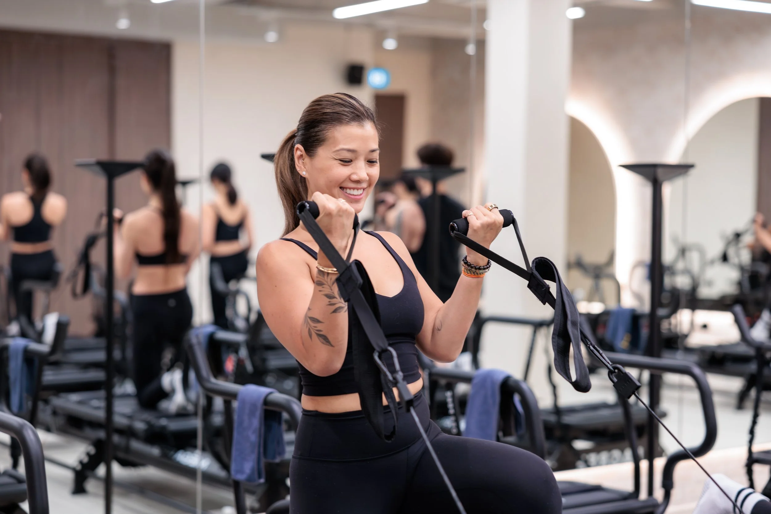 Young woman in a black sports bra and leggings uses a resistance band in a gym. She is smiling, surrounded by workout equipment and mirror reflections. HKore Megaformer Lagree Yoga Studio Photography
