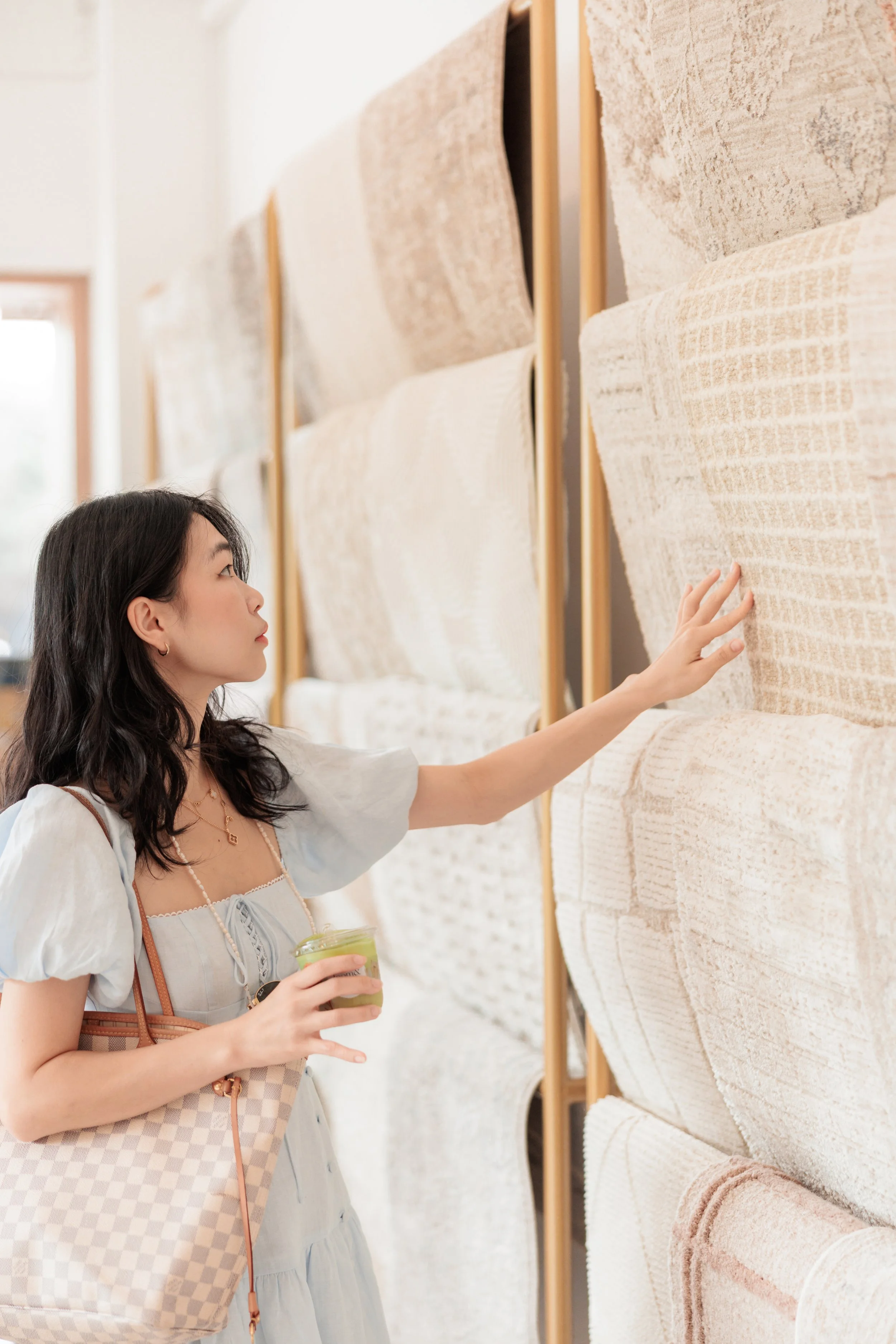 A woman with dark hair and a checkered bag looks at and touches a textured beige and white rug on display in a store or showroom. Loom & Stone Store Opening. Event Photography. Event Videography. Rugs. Carpets. Marimekko rugs.