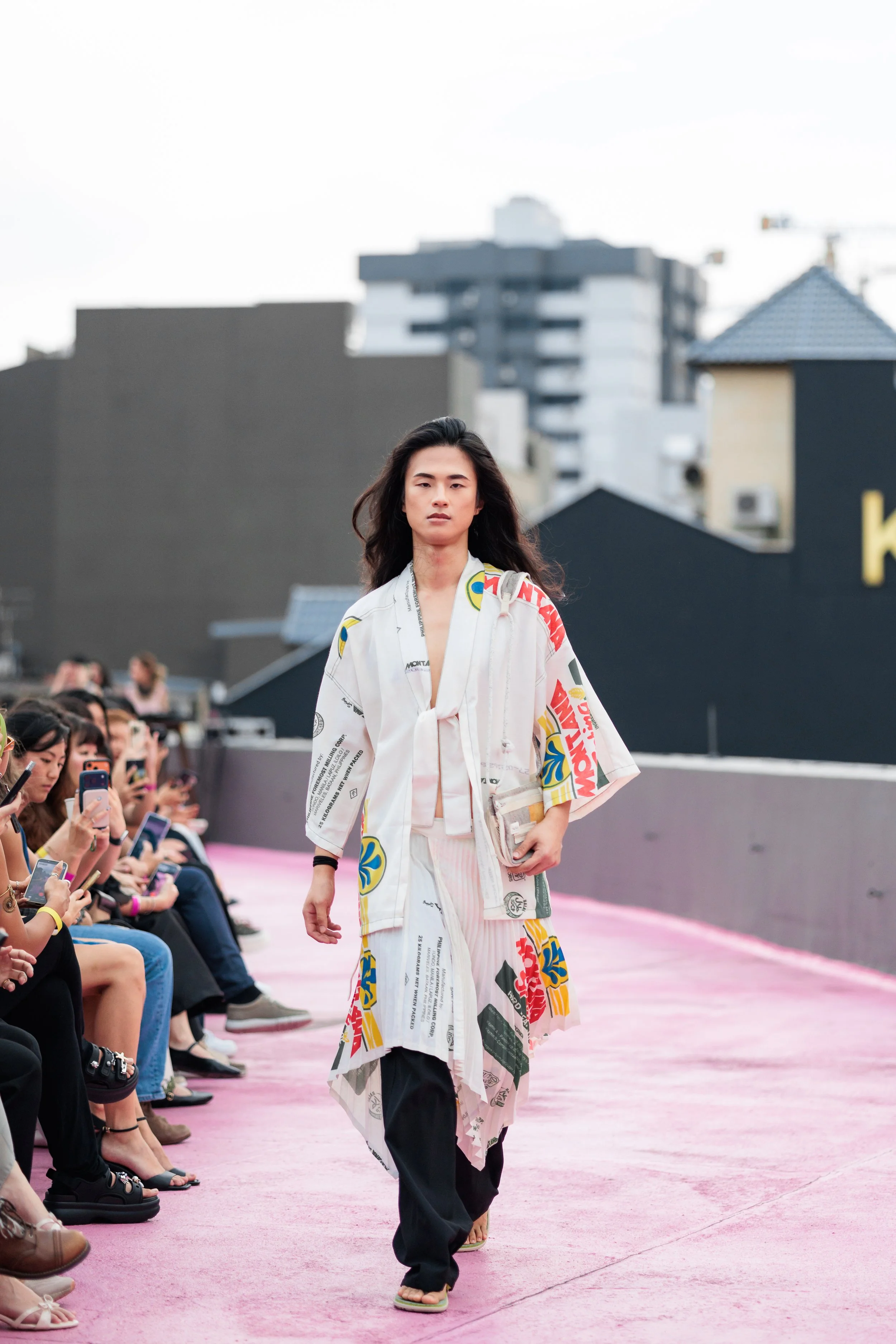 A woman walking on a pink runway during a fashion show, wearing a white coat and pants with colorful patches and text, and holding a clutch bag. Event Photography. Fashion Photography. Event Videography. Walkway Photography & Videography.
