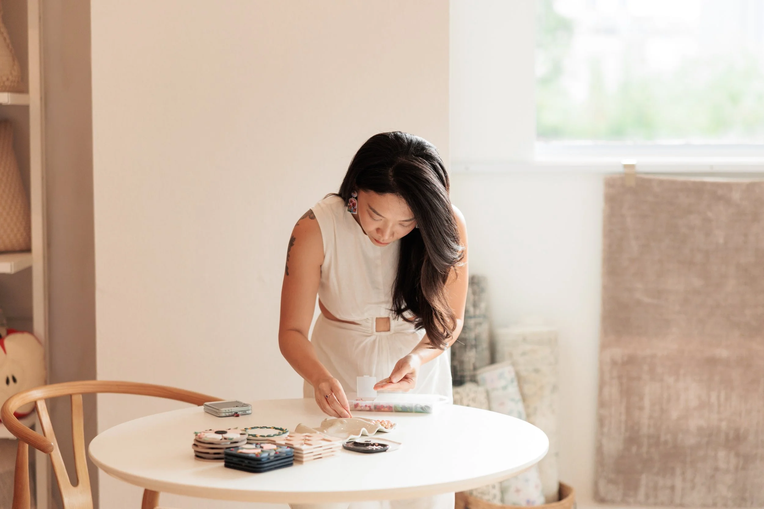A woman with long dark hair stands at a round white table, looking down while working on a craft project with holiday-themed ornaments and decorations, in a bright, cozy room. Loom & Stone Store Opening. Event Photography. Event Videography. Rugs.