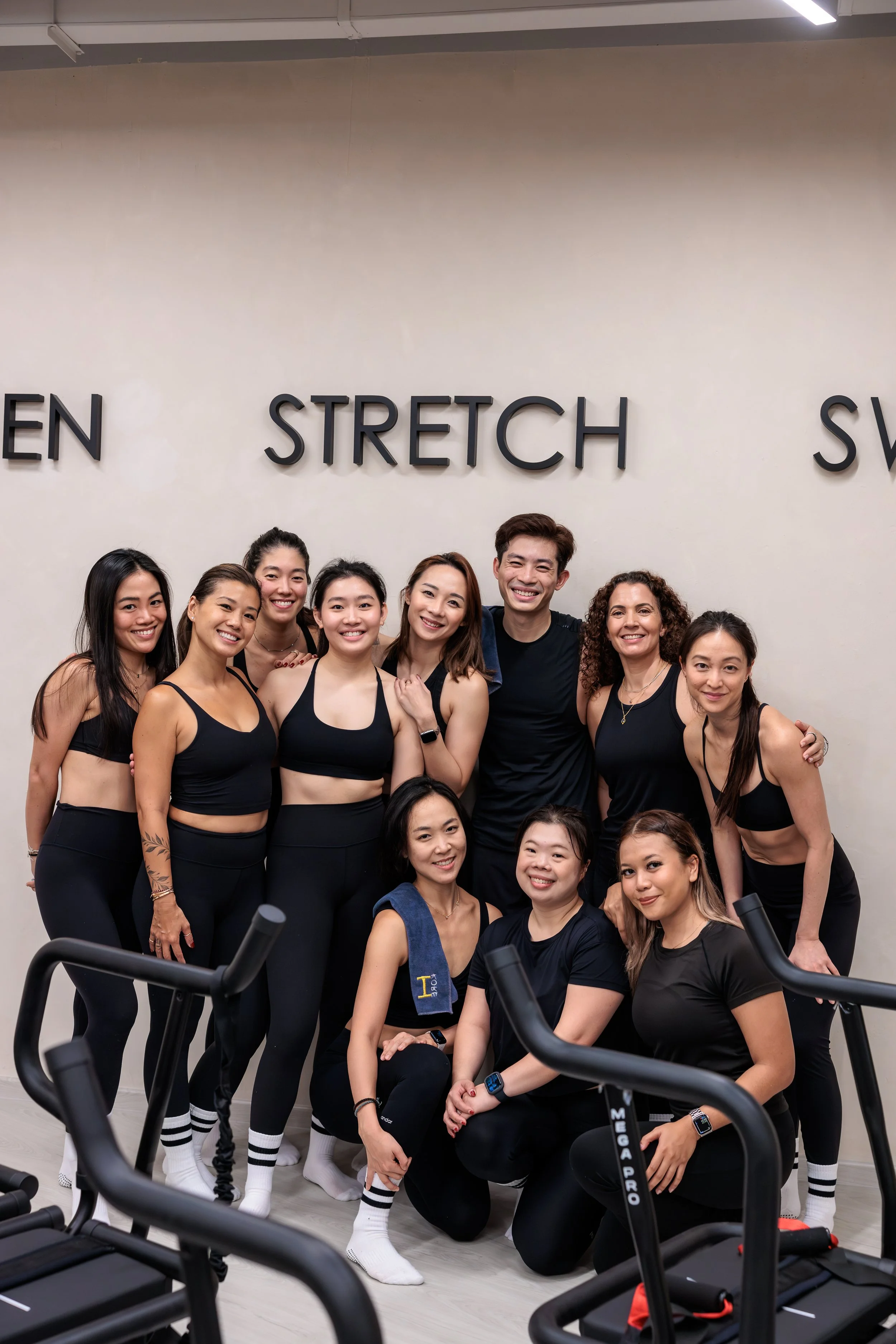 Group of eight women and one man in workout clothes posing for a photo in a fitness studio, with the words 'STRETCH' and 'SW' on the wall behind them. HKore Megaformer Lagree Yoga Studio Photography