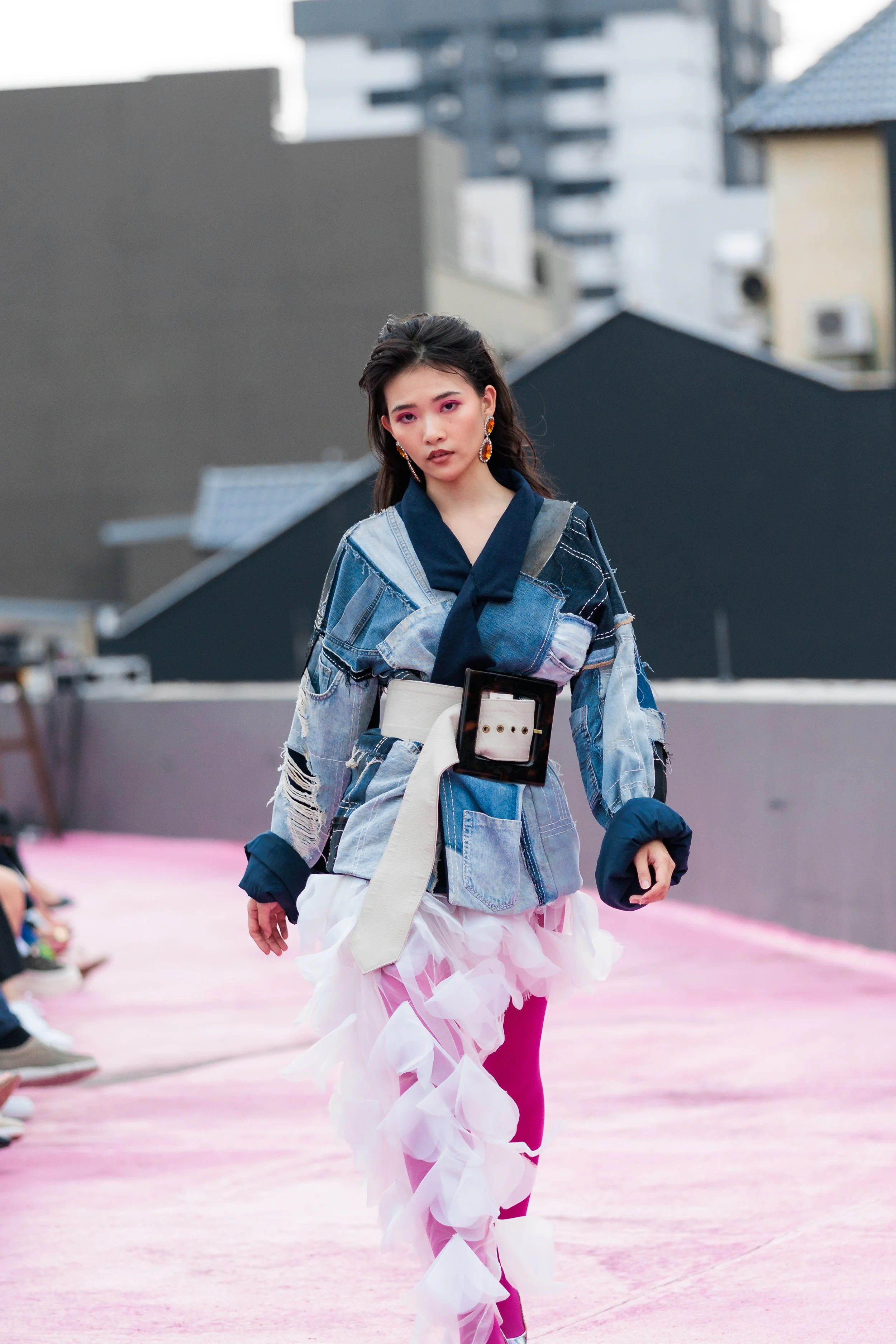 A female model walking on a pink runway during a fashion show, wearing a patchwork denim jacket with a black scarf, a white belt with a large buckle, and a fluffy pink skirt with ruffles. She has long dark hair, bold makeup with pink eyeshadow, and c