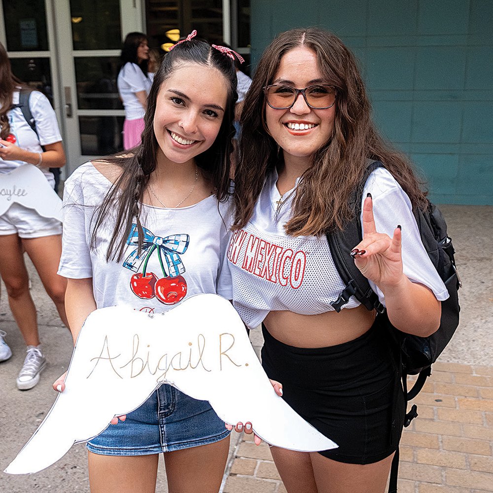Two young women smiling, holding a sign with the name Abigail R, at an outdoor event. One woman wears a white t-shirt with cherry graphics, the other wears glasses and a white shirt with red lettering. In the background, other young women are visible.