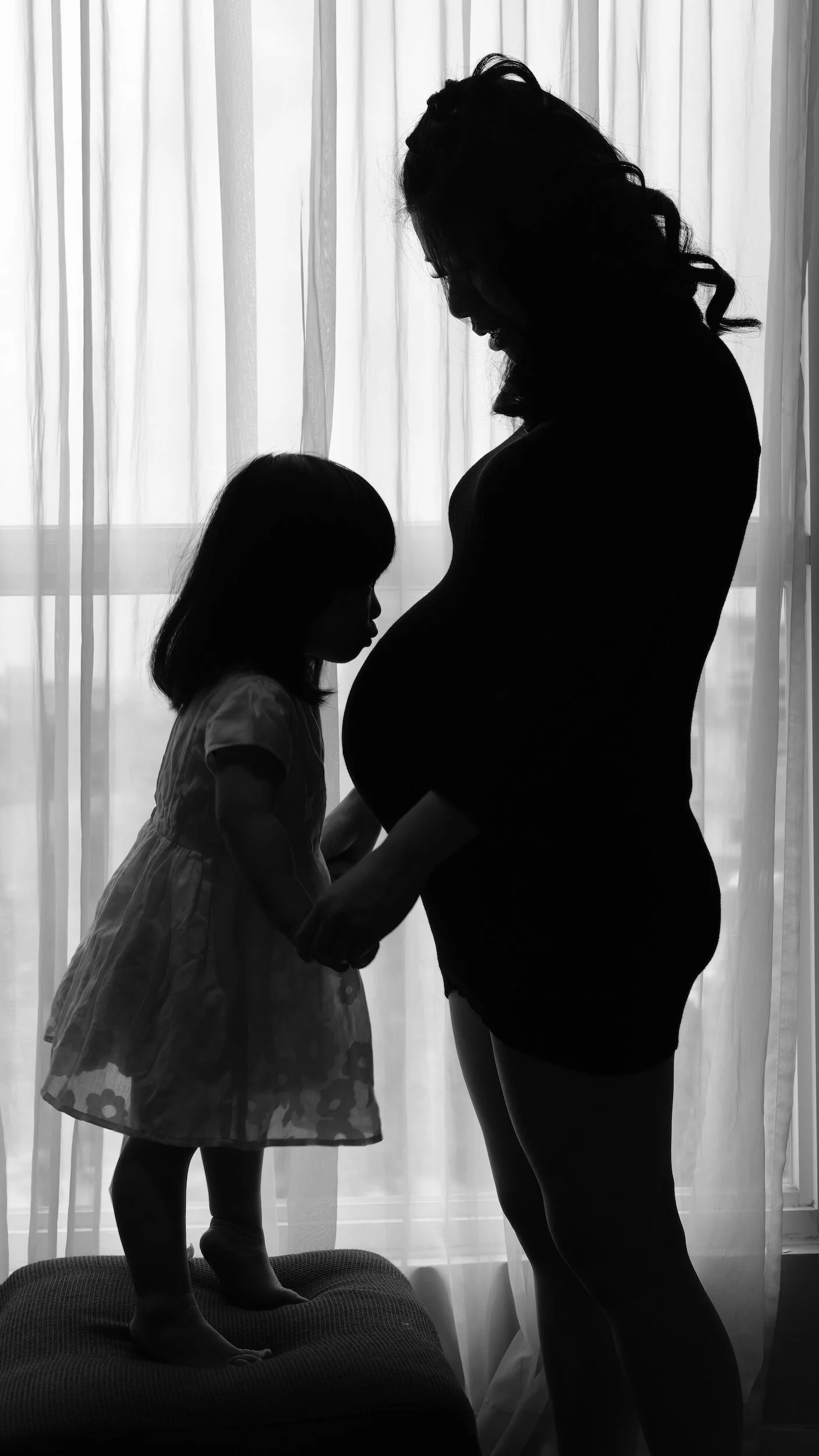Silhouette of a pregnant woman and a young girl holding hands in front of a window with light curtains.