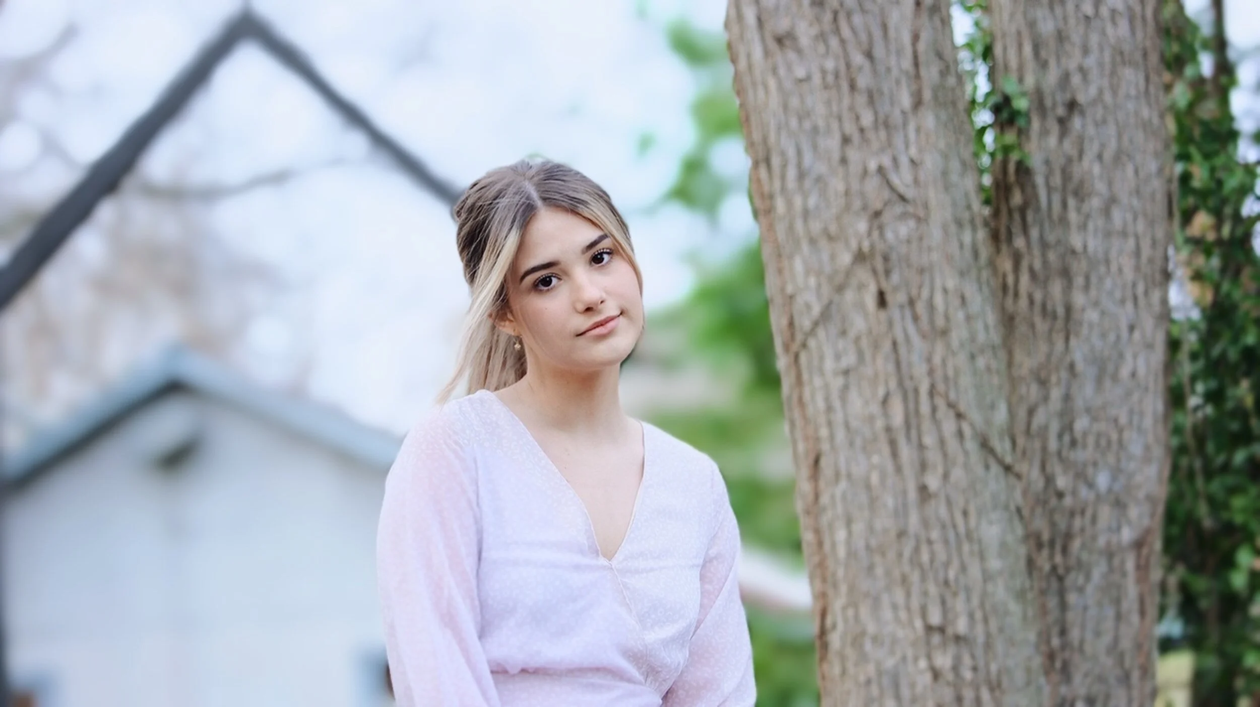 A young woman with blonde hair and light skin standing outdoors next to a tree, wearing a light pink blouse, with a neutral expression on her face.