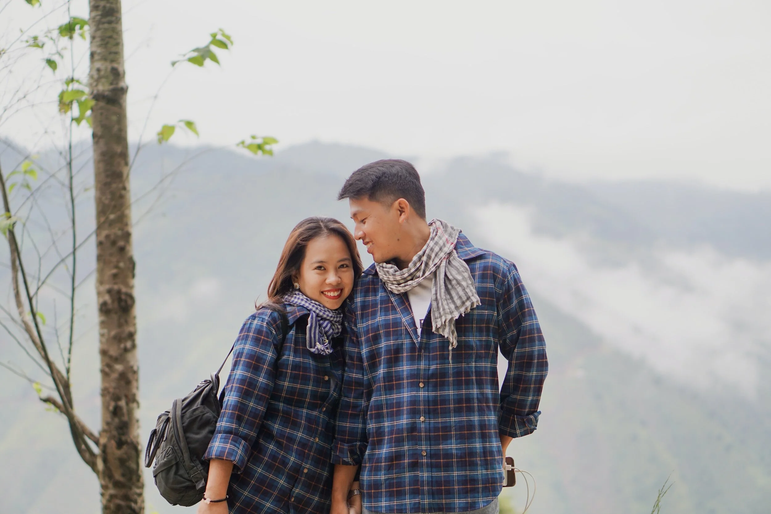 Smiling young woman and man in plaid shirts sharing a moment outdoors in a misty mountain landscape.