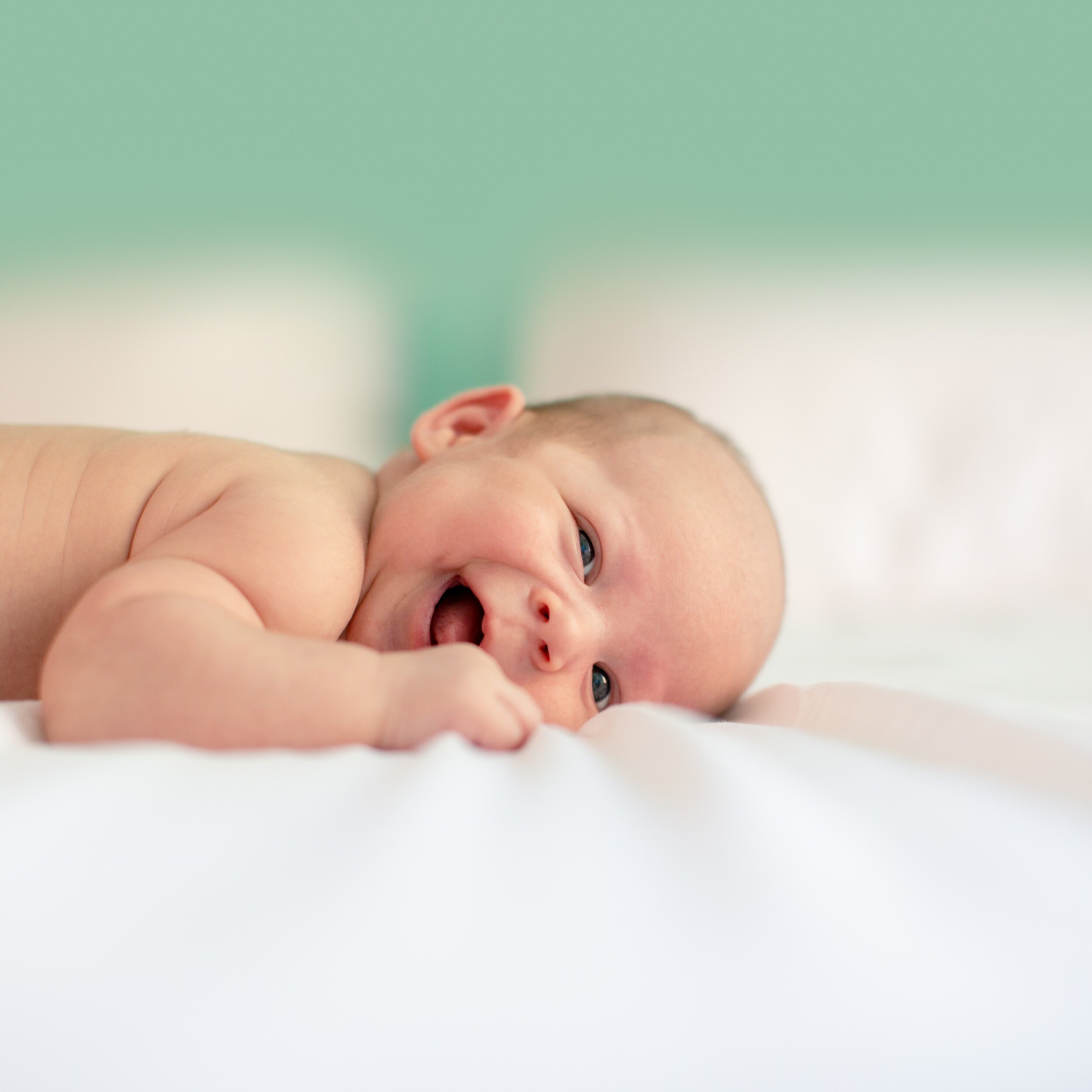 A newborn baby lying on a white surface, looking at the camera with a slightly open mouth and bright eyes, with a green and white background.