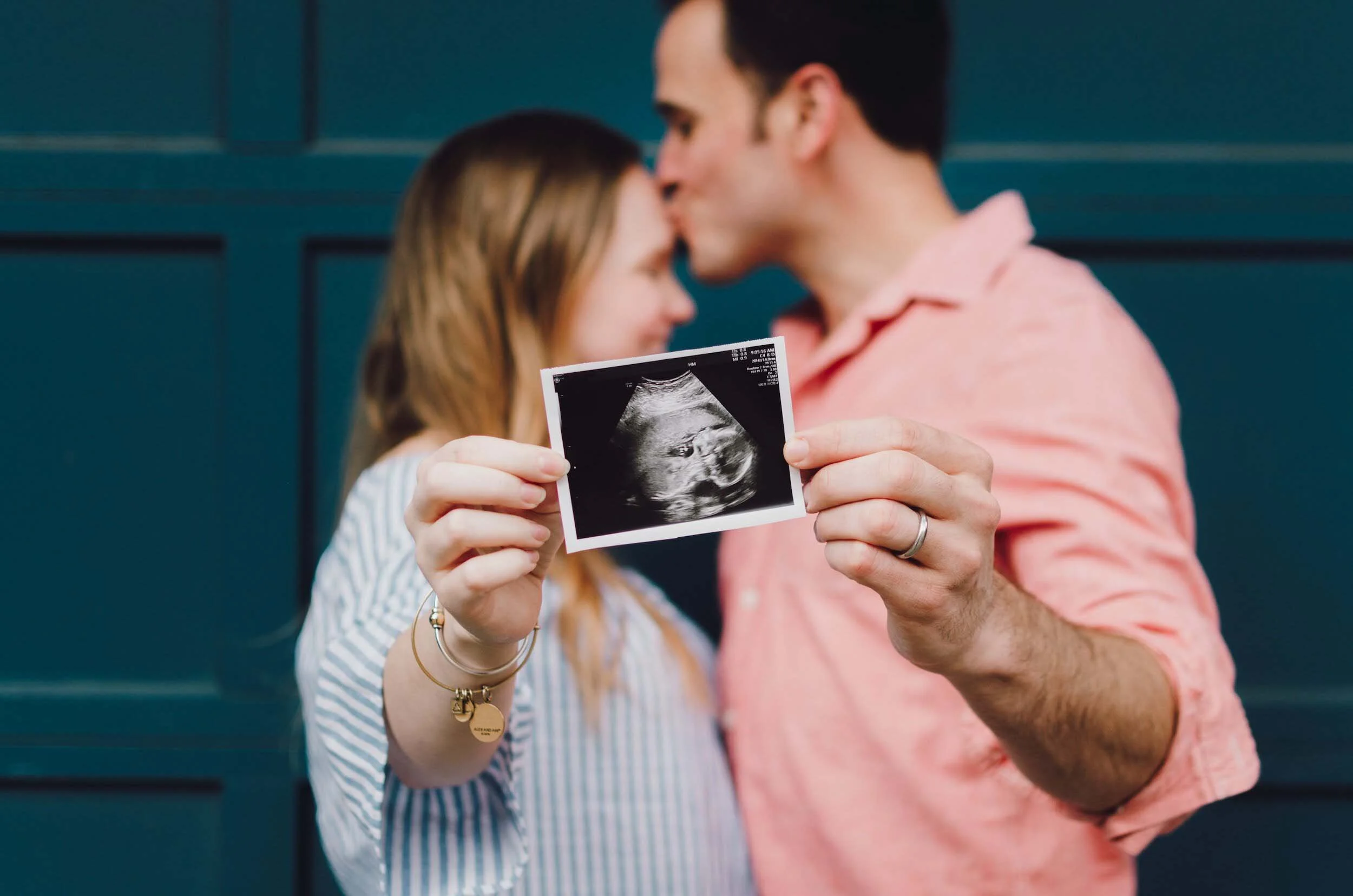 A couple holding an ultrasound image of a baby, with the man kissing the woman on the forehead.