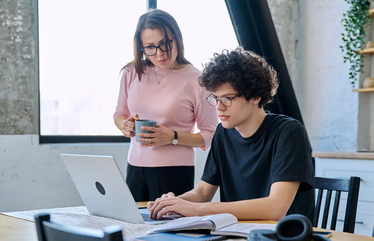 Mother and son college student at kitchen table with laptop reviewing documents, mother holding cup of coffee