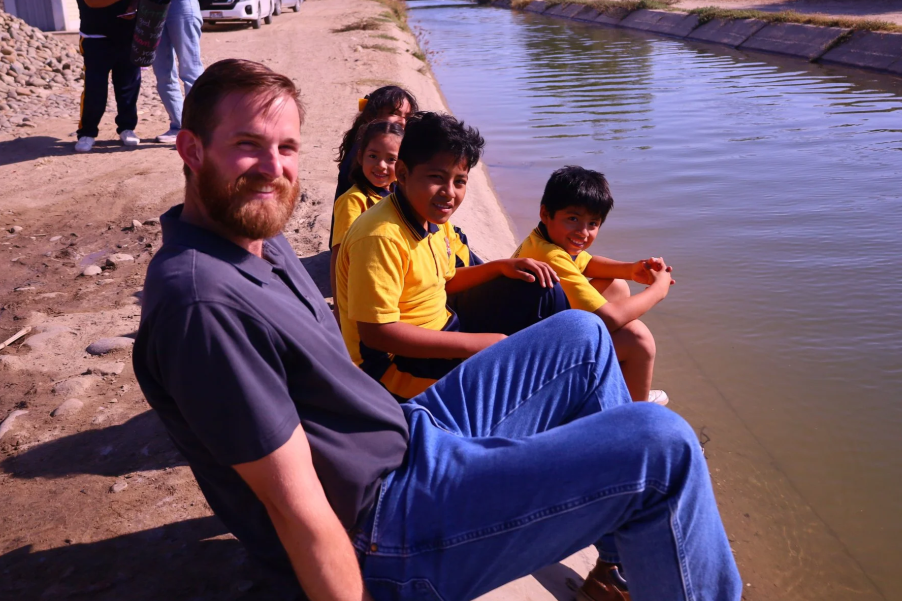 A group of people sitting by the edge of a body of water, with smiling children in yellow shirts and an adult man in a dark shirt in the foreground, and a few more people standing behind them.