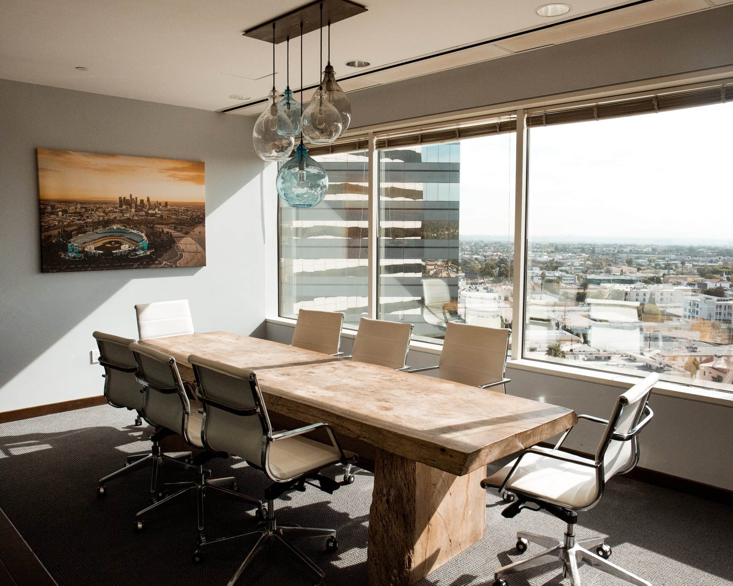 Conference room with with a wooden desk, tan chairs and a large window bathing the room in sunlight