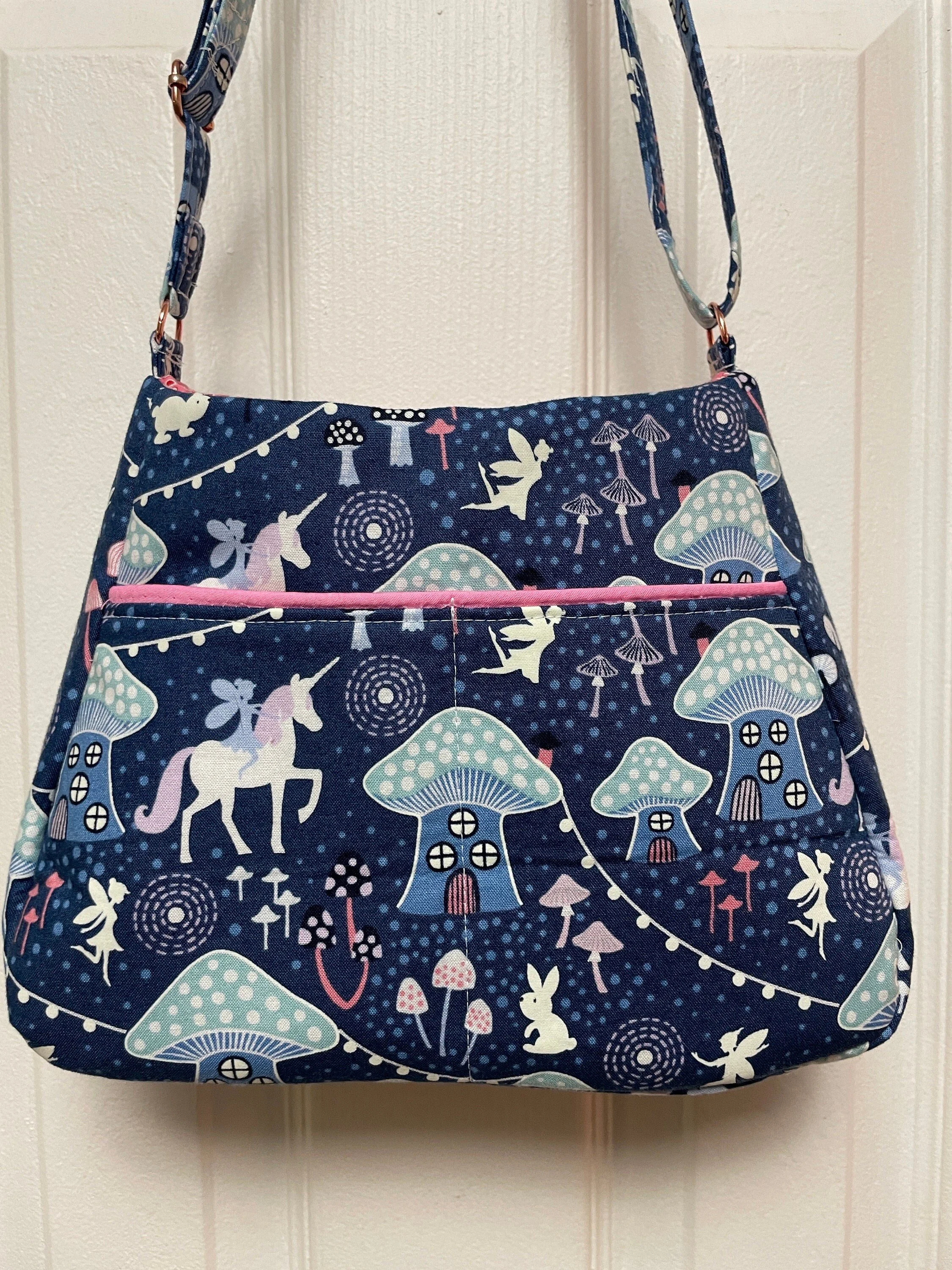 A fabric shoulder bag hanging on a white door, featuring a whimsical pattern with unicorns, rabbits, mushrooms, fairy tale houses, and magical elements in pastel colors on a dark blue background.