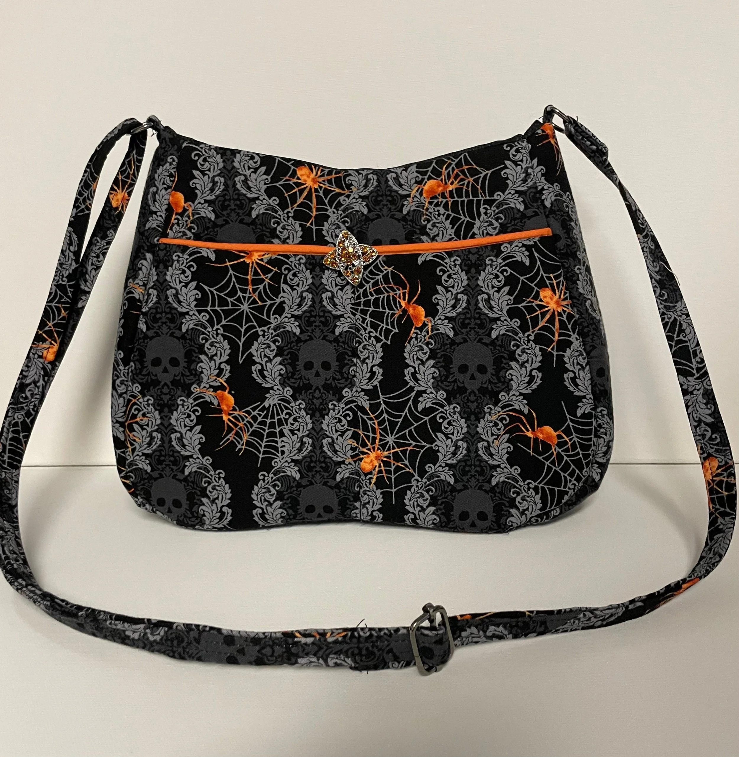 Black shoulder bag with orange spiderweb and skull pattern, orange zipper, and a decorative pin on the front.