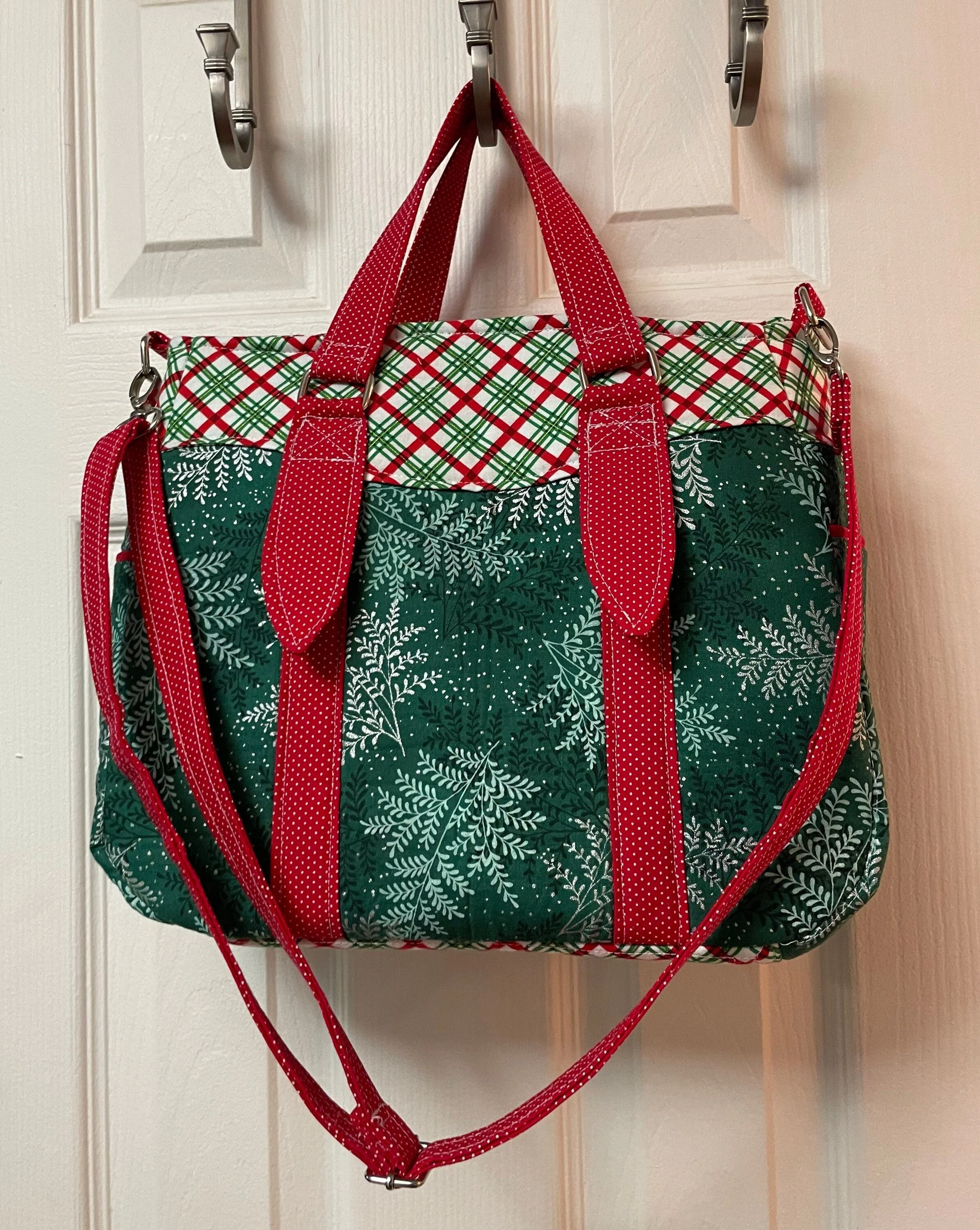 A Christmas-themed tote bag hanging on a door with a white background. The bag features green, white, and red patterns, including snowflakes and plaid, with red straps and handles.