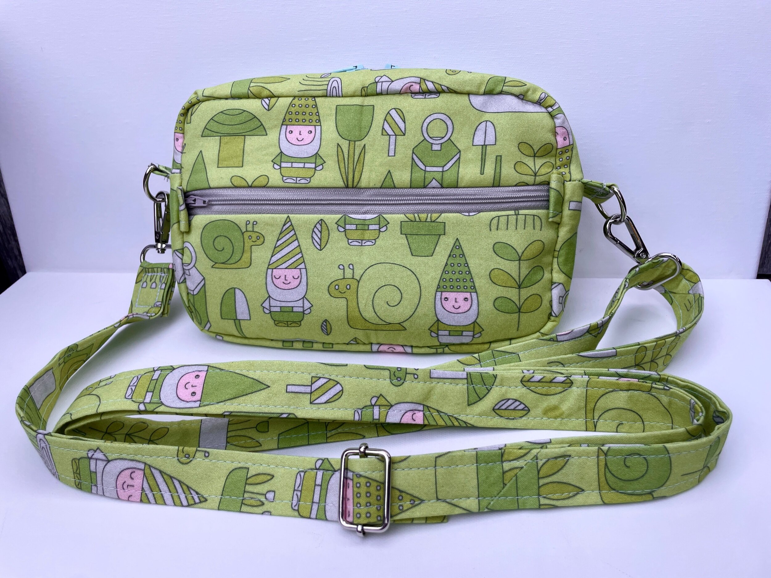 A light green fabric bag with a cartoon garden pattern, including snails, snails, gnomes, mushrooms, and plants. It features a zippered pocket on the front and a long adjustable strap with a metal buckle.