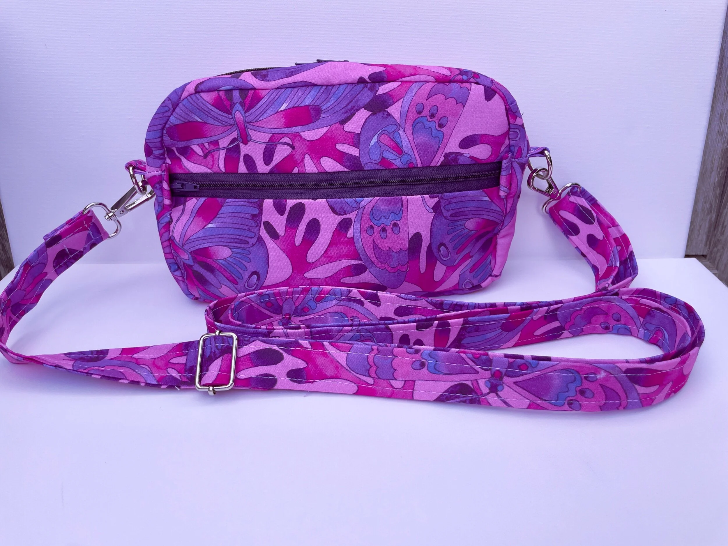 Pink and purple floral patterned crossbody bag with adjustable strap and front zipper pocket.