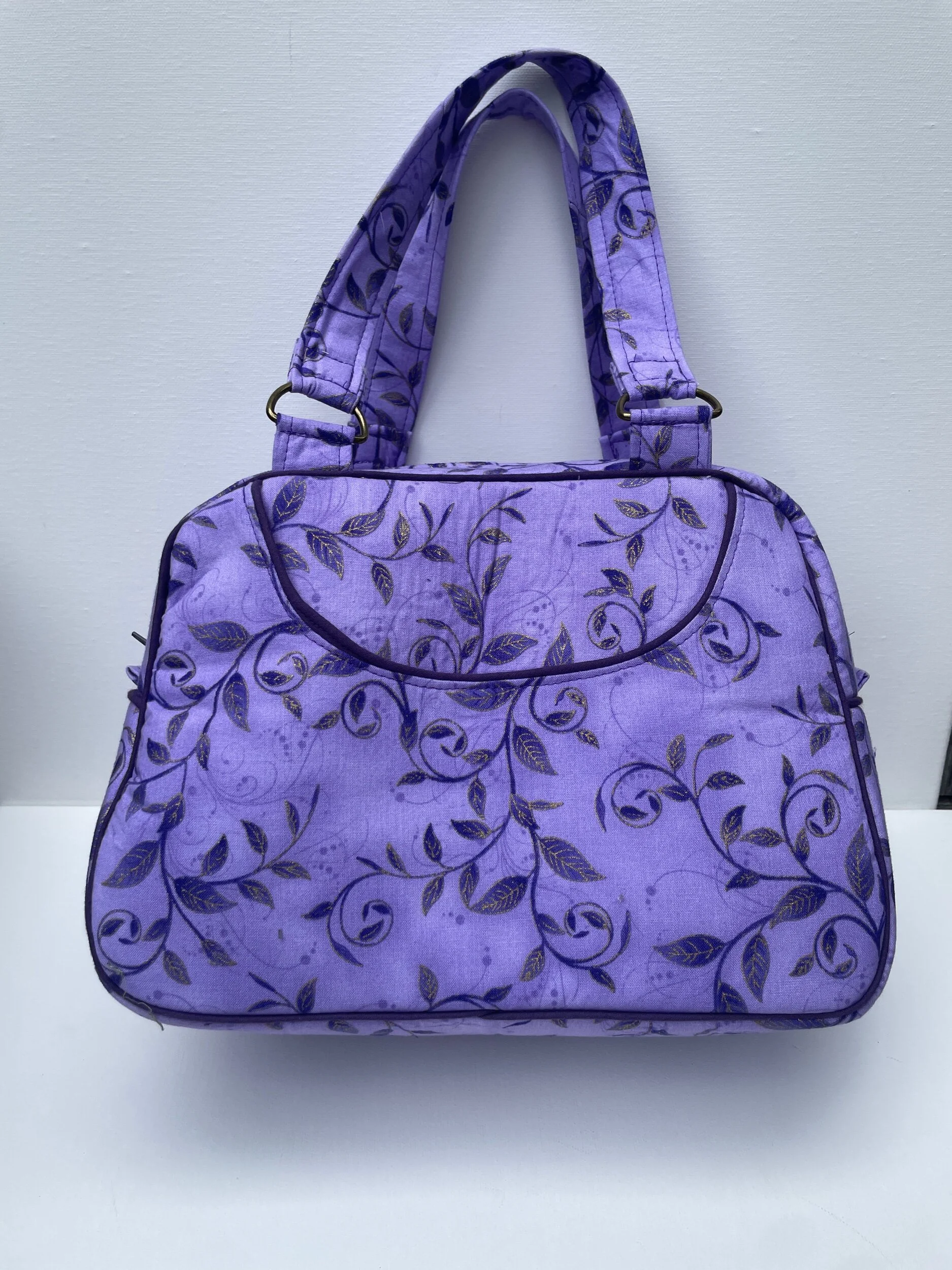 Purple handbag with a floral pattern, handles on top, placed against a white wall.