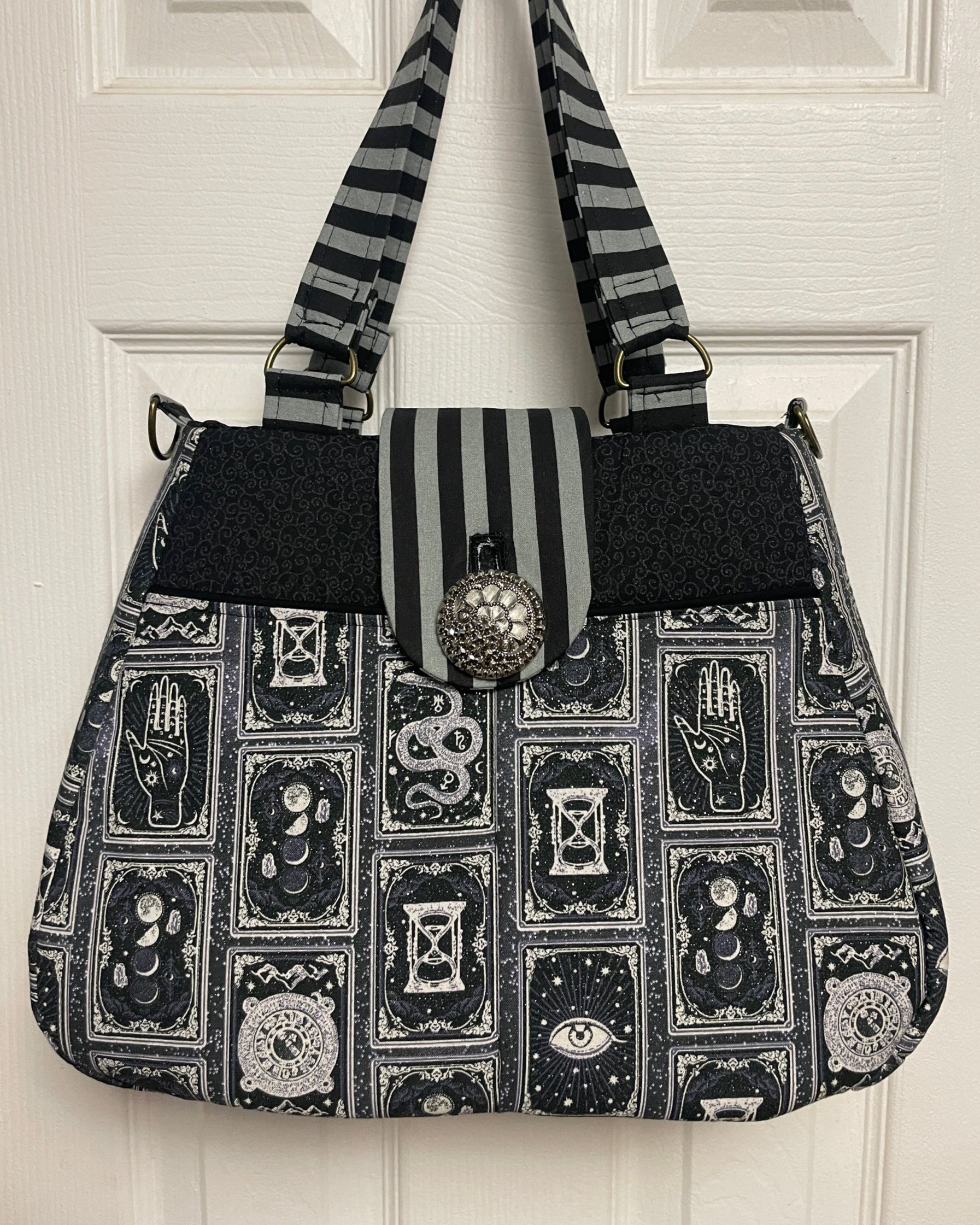 A black and white patterned fabric bag with tarot card designs, decorative clasp, and striped straps hanging on a white door.