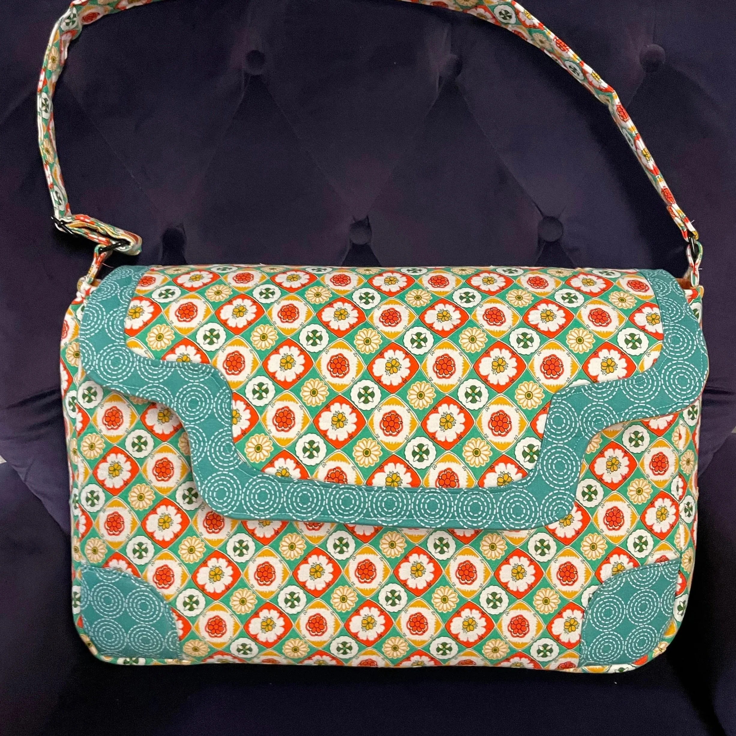 Colorful patterned shoulder bag with floral and geometric designs in orange, white, yellow, green, and blue, resting on a dark surface.